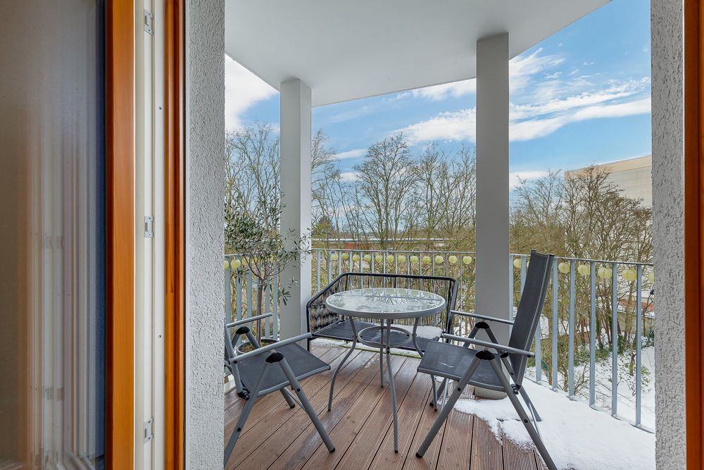 Balkon mit Holzboden, zwei Stühlen und einem runden Glastisch, Blick auf Bäume.