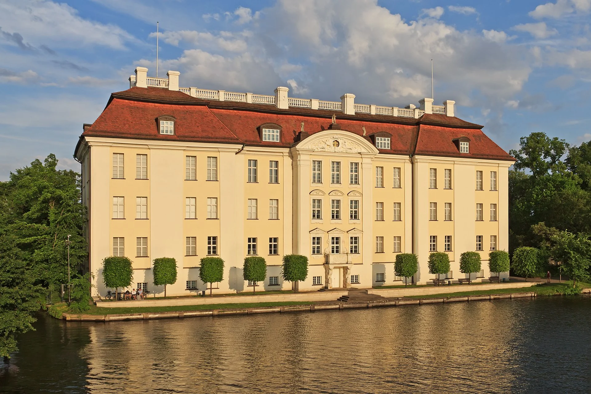 Großes, helles Wohngebäude mit rotem Dach, vielen Fenstern und Bäumen am Wasser.