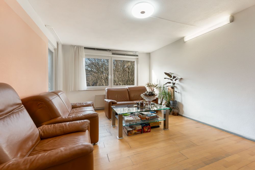 Wohnzimmer mit braunen Ledersofas, Glastisch und Pflanzen am Fenster.