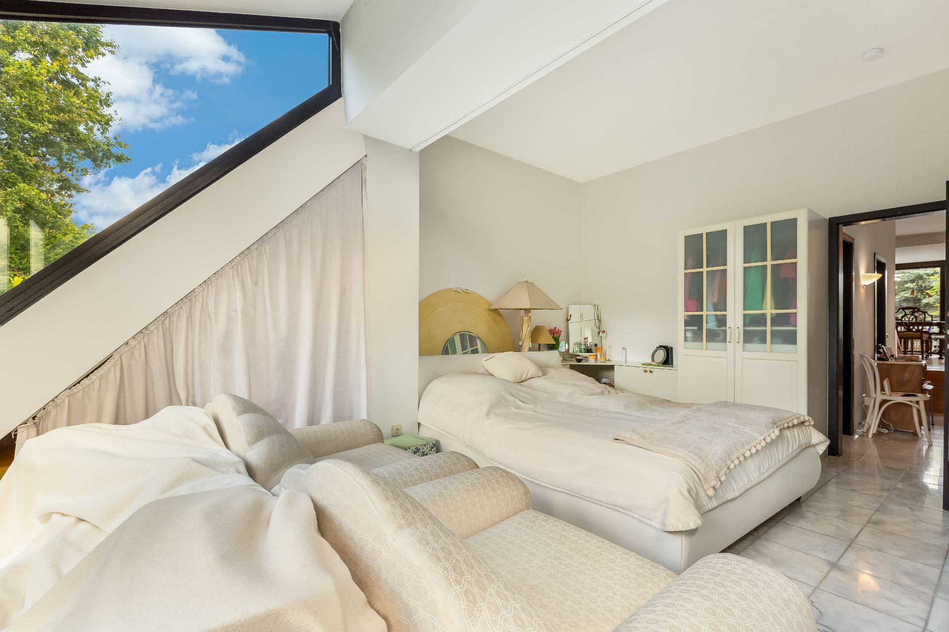 Helles Schlafzimmer mit großem Fenster, Bett, Sofa und Schrank auf Fliesenboden.