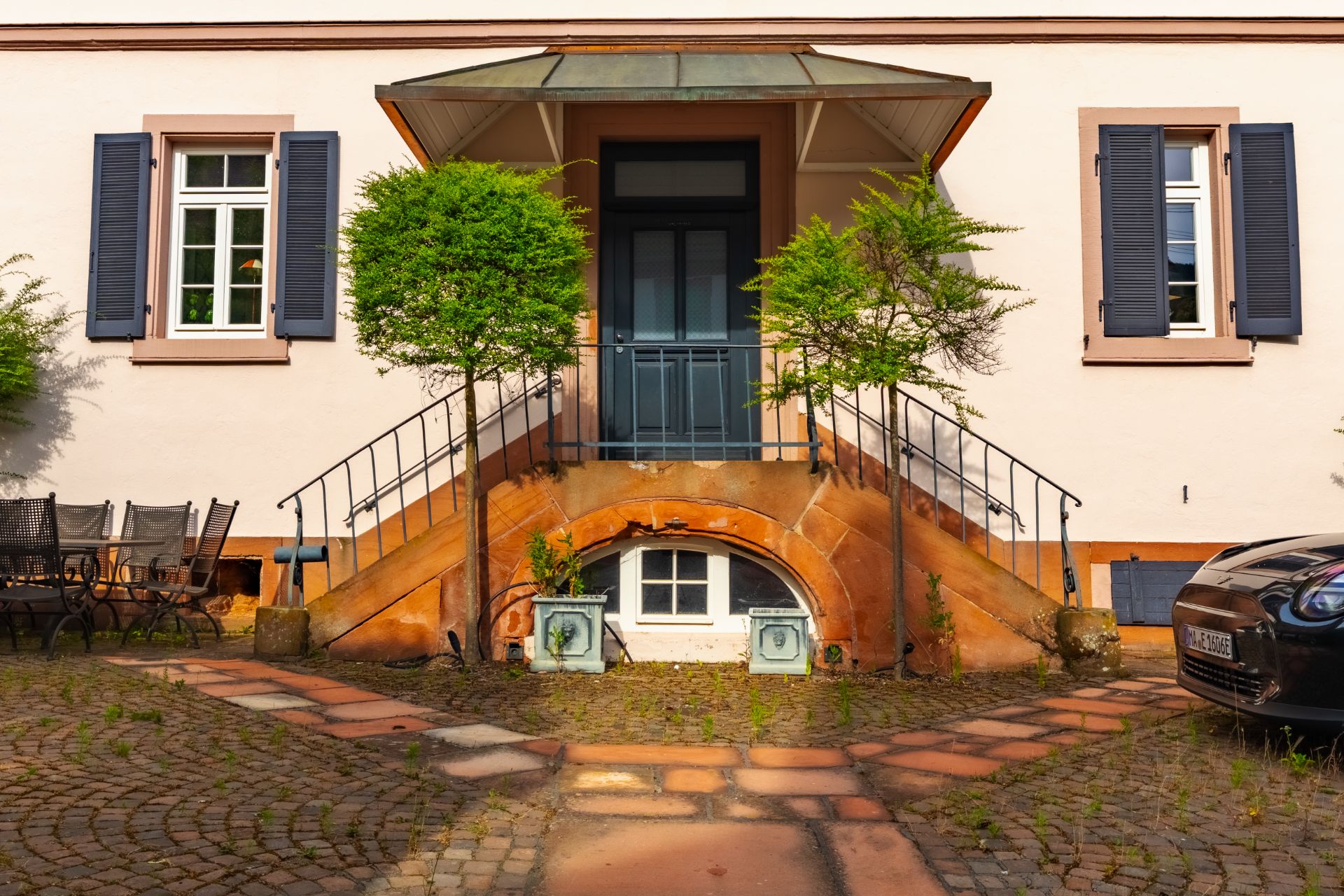 Eingang mit Doppeltreppe, zwei Fenster mit blauen Klappläden, davor Kopfsteinpflaster.