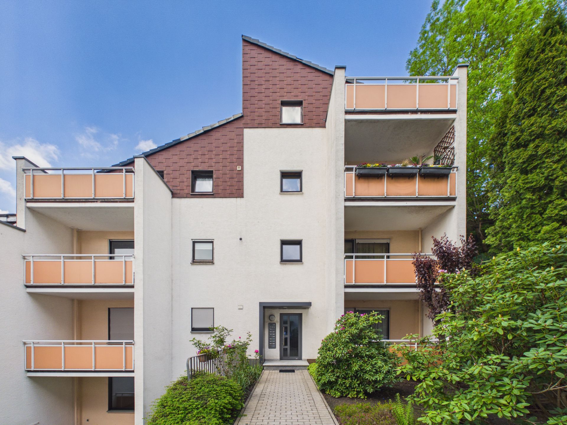Modernes Mehrfamilienhaus mit Balkonen, helle Fassade und Eingang, umgeben von Pflanzen.