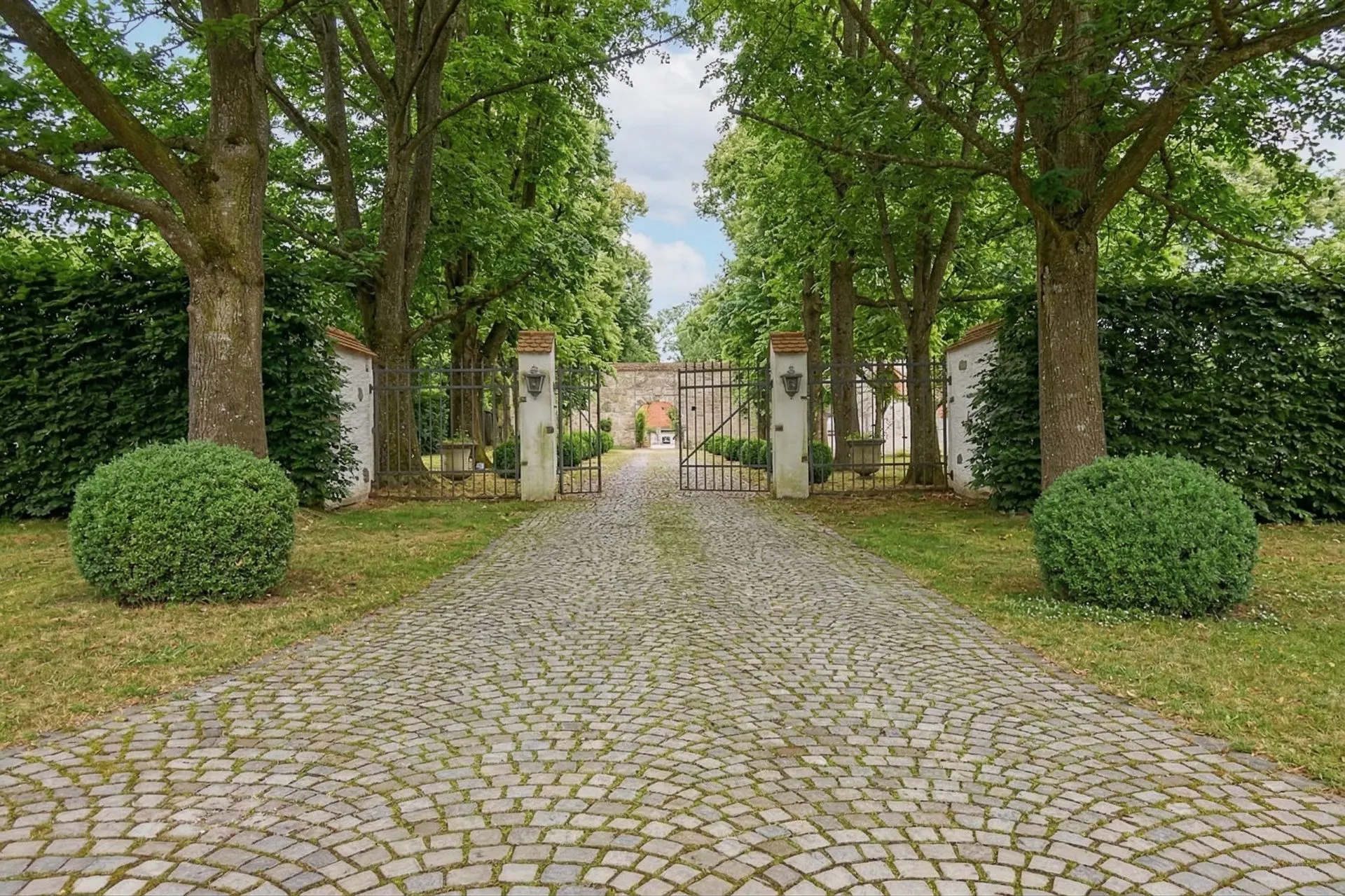 Kopfsteinpflasterweg führt durch ein Tor, flankiert von Bäumen und Kugelsträuchern.