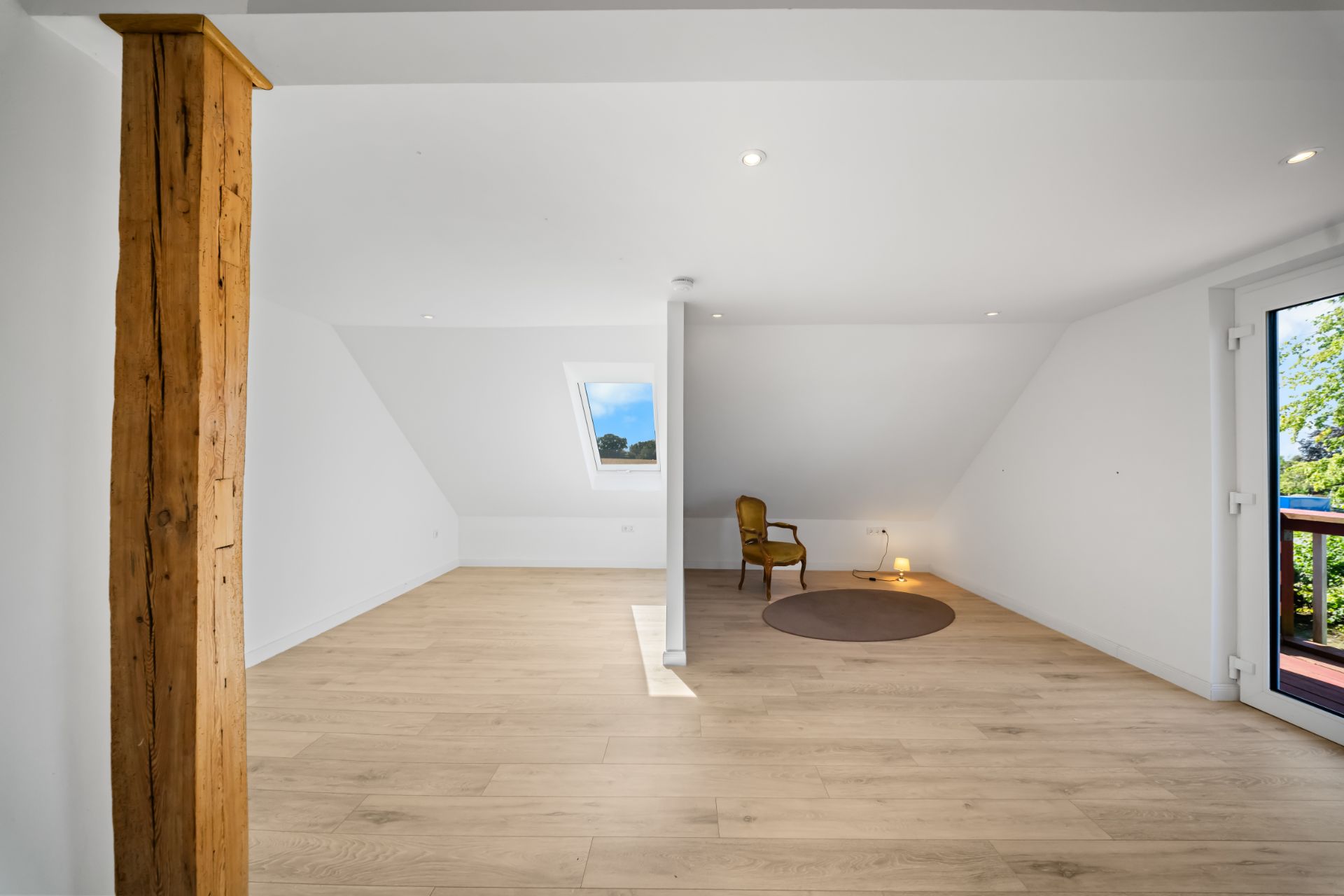 Helles Dachzimmer mit Holzfußboden, Stützbalken, Sessel und Fenstertür zum Balkon.