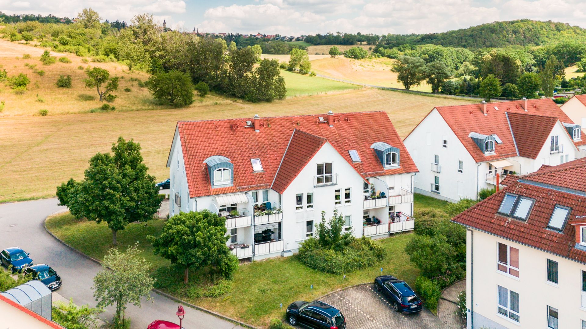 Mehrfamilienhaus mit rotem Dach, Balkonen und parkenden Autos, umgeben von Bäumen und Wiesen.