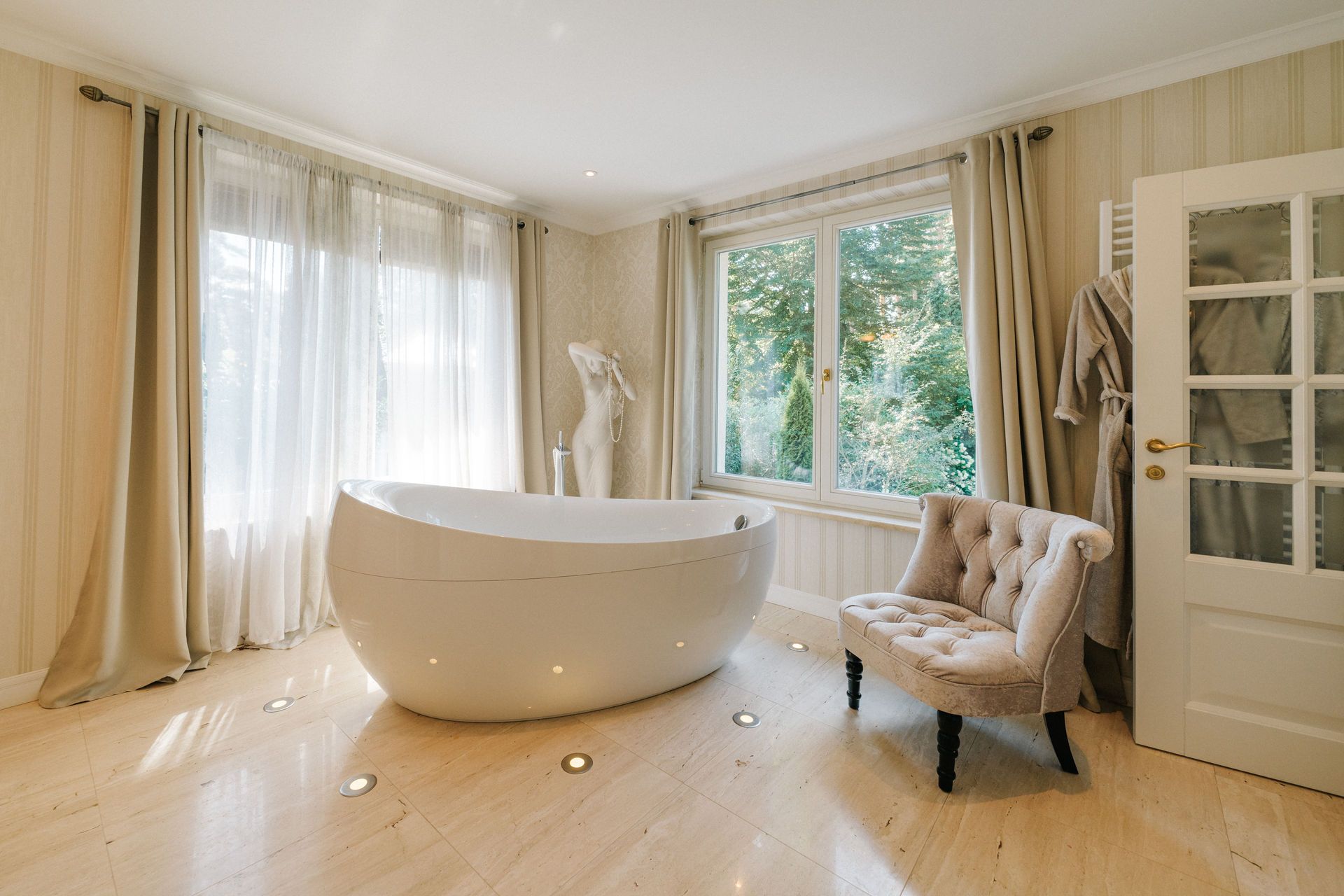 Helles Badezimmer mit freistehender Badewanne, Sessel, großen Fenstern und Vorhängen.