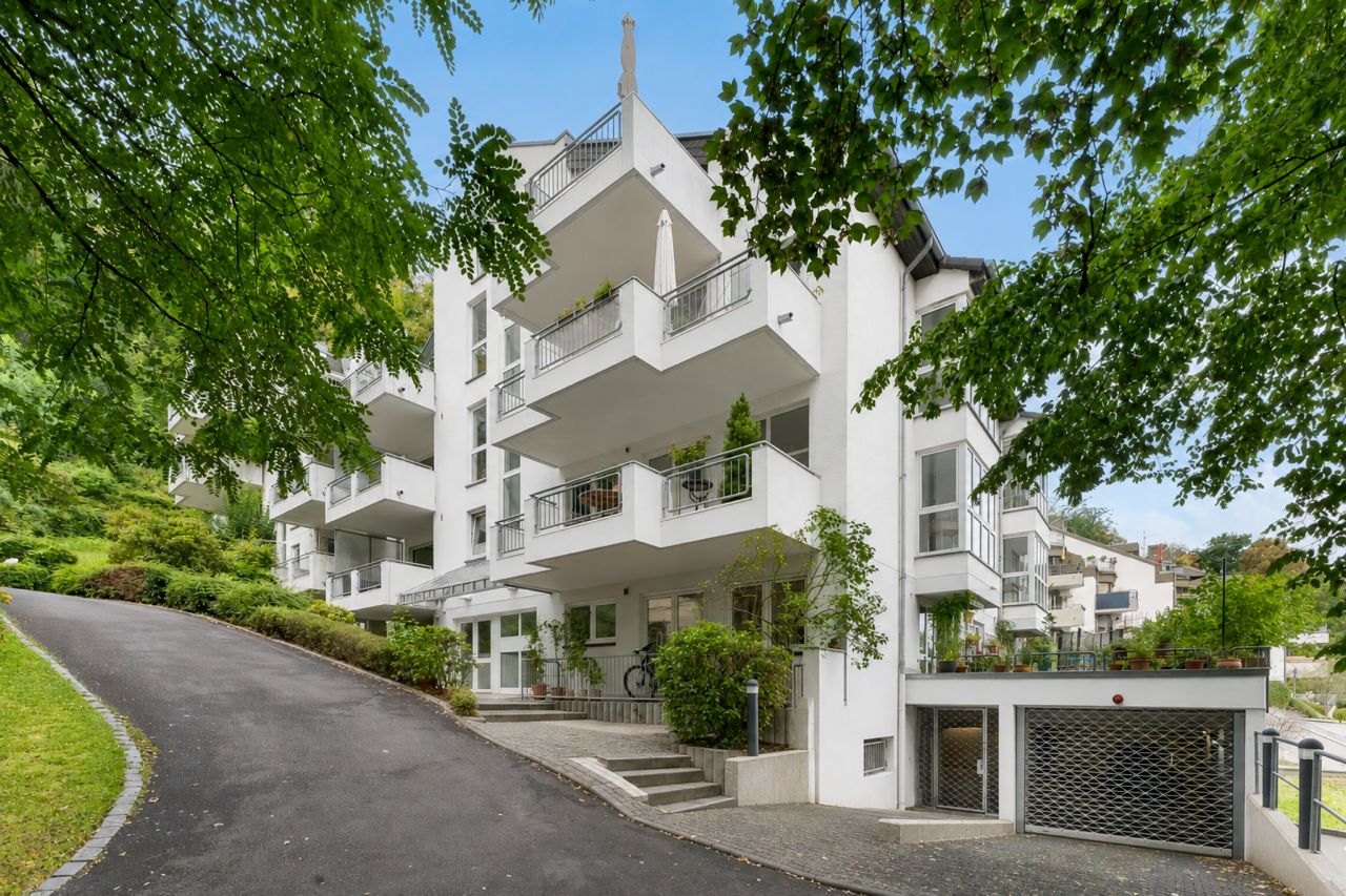 Modernes, weißes Mehrfamilienhaus mit mehreren Balkonen und grünen Pflanzen vor dem Eingang.