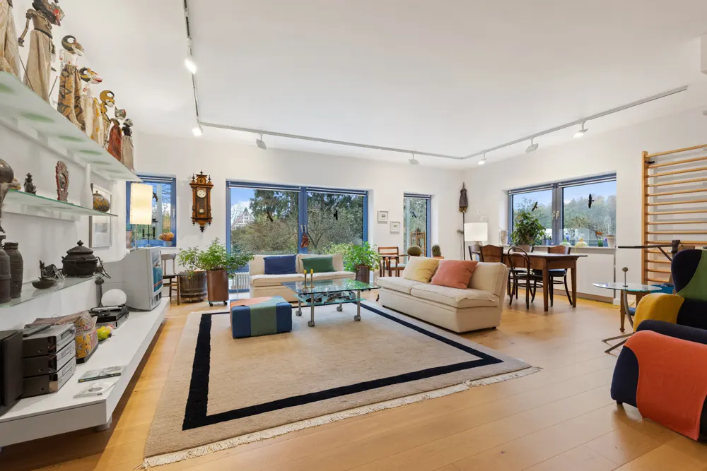 Modernes Wohnzimmer mit großen Fenstern, Sofa, Teppich, Esstisch und Wandregalen mit Dekoration.