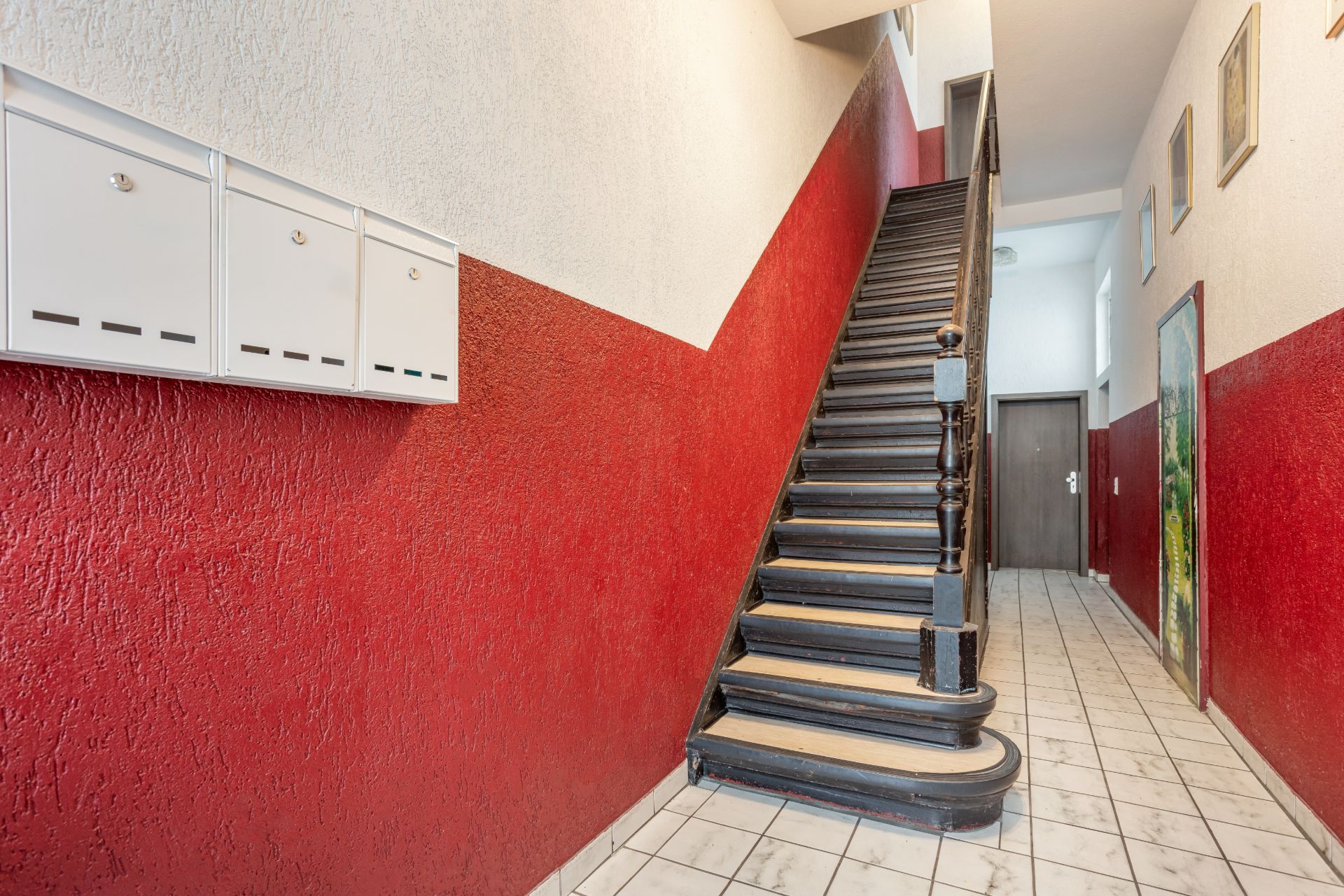 Treppenhaus mit roter Wand, Briefkästen und Fliesenboden in einem Wohnhaus.