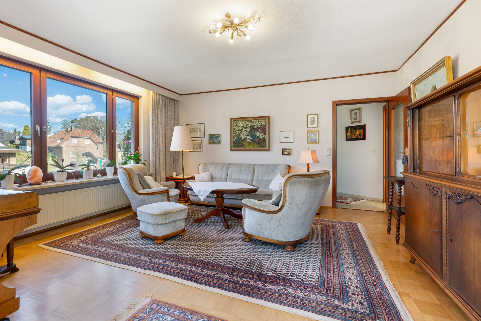 Helles Wohnzimmer mit Sitzgruppe, großem Fenster, Teppich und mehreren Bildern an den Wänden.