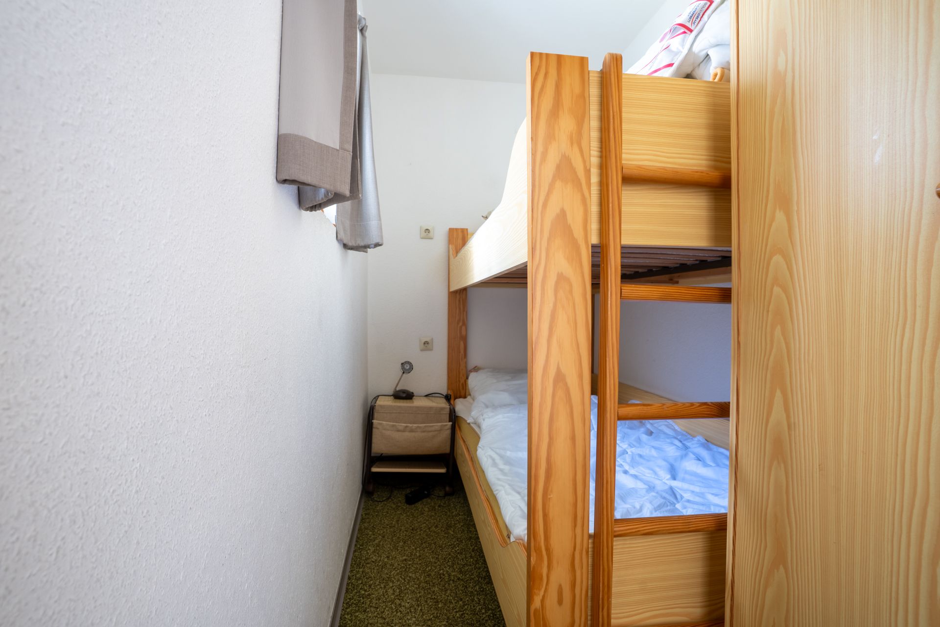 Schmales Zimmer mit einem Etagenbett aus Holz und einem kleinen Nachttisch am Fenster.