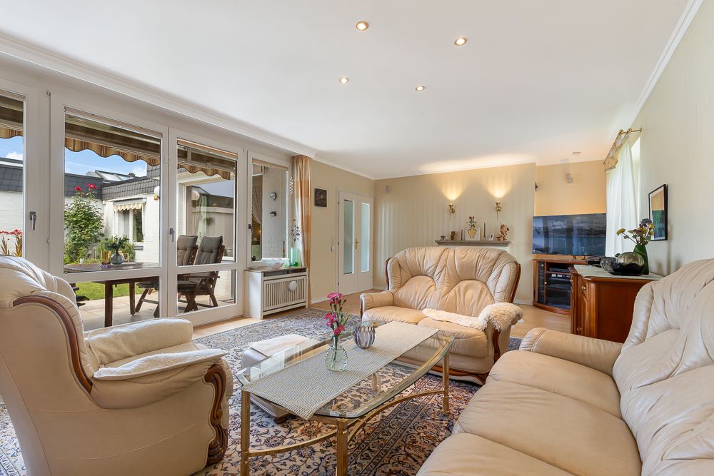 Helles Wohnzimmer mit beigen Ledersofas, Glastisch und großen Fenstern zum Garten.
