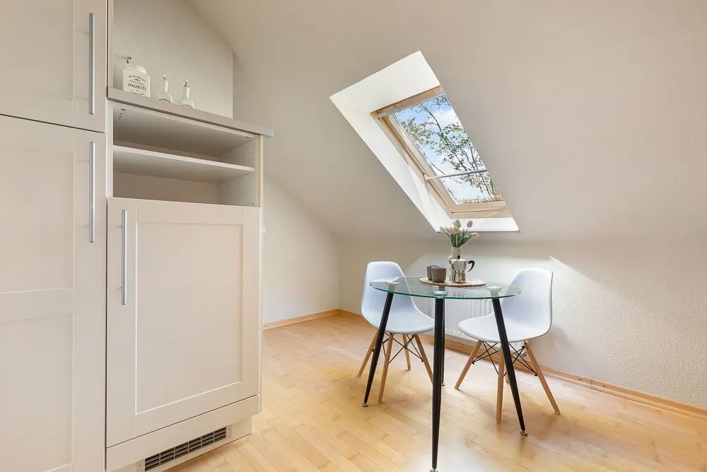 Heller Raum mit Dachfenster, kleinem runden Glastisch und zwei weißen Stühlen auf Holzfußboden.
