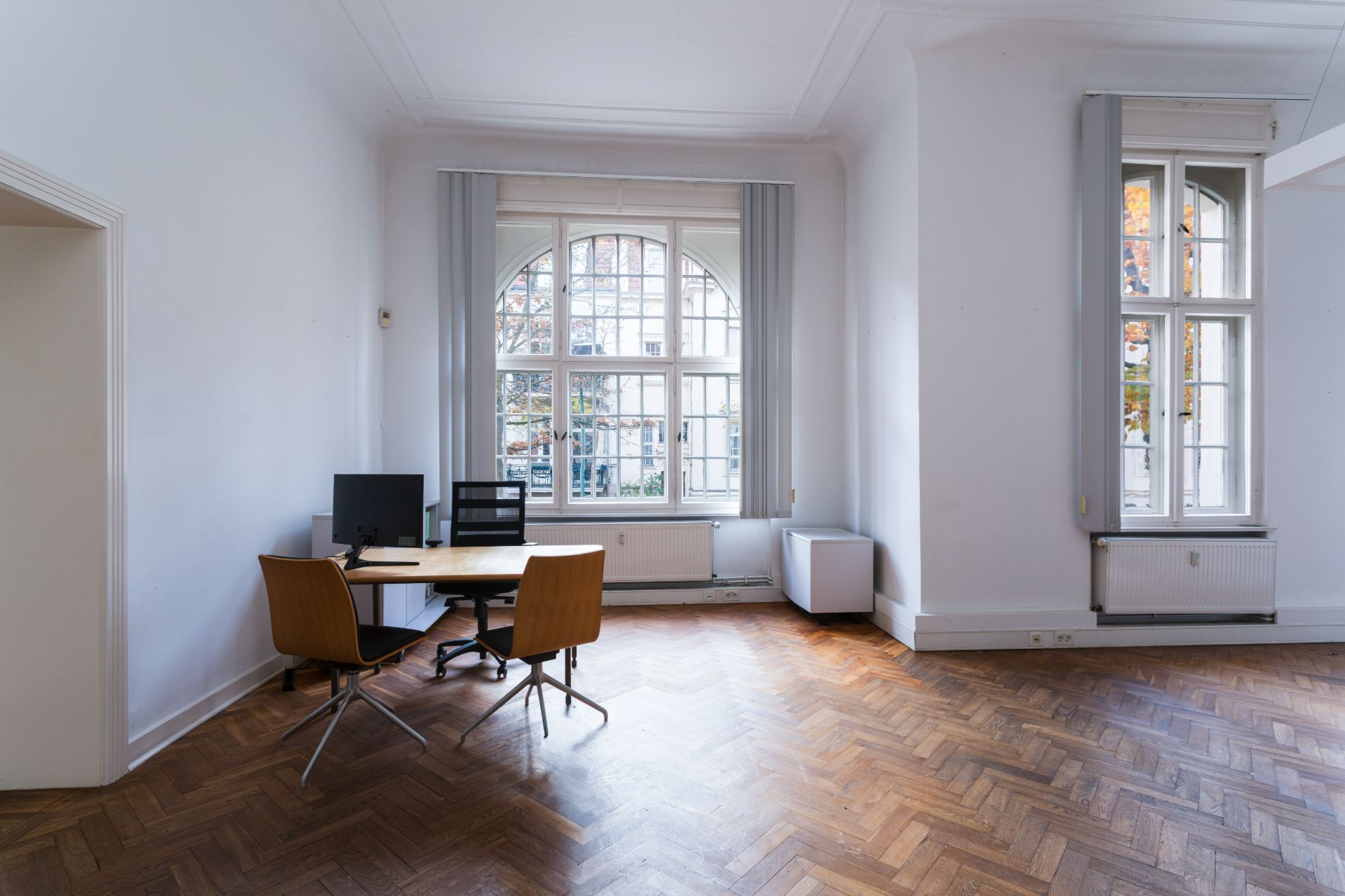 Helles Zimmer mit großem Fenster, Parkettboden und Schreibtisch mit Computer und zwei Stühlen.