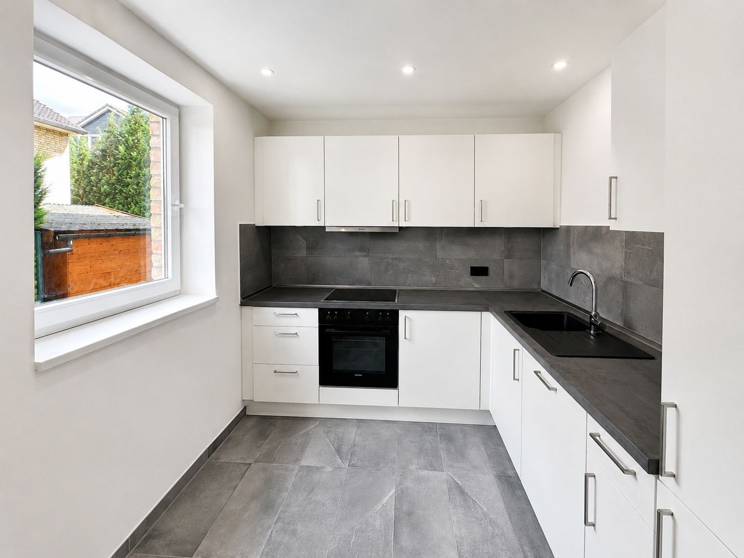 Moderne Küche mit weißen Schränken, grauen Fliesen und großem Fenster.