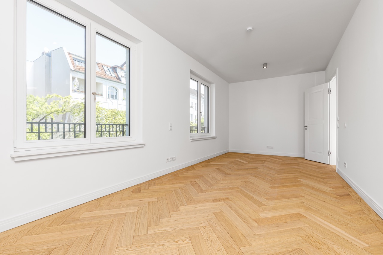 Helles, leeres Zimmer mit weißer Wand, Parkettboden und zwei großen Fenstern.