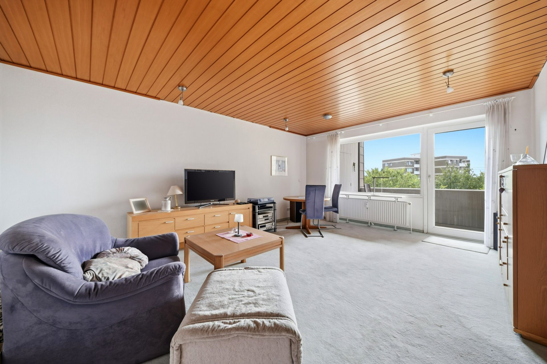 Helles Wohnzimmer mit Sessel, Fernseher, Schreibtisch und Zugang zum Balkon.