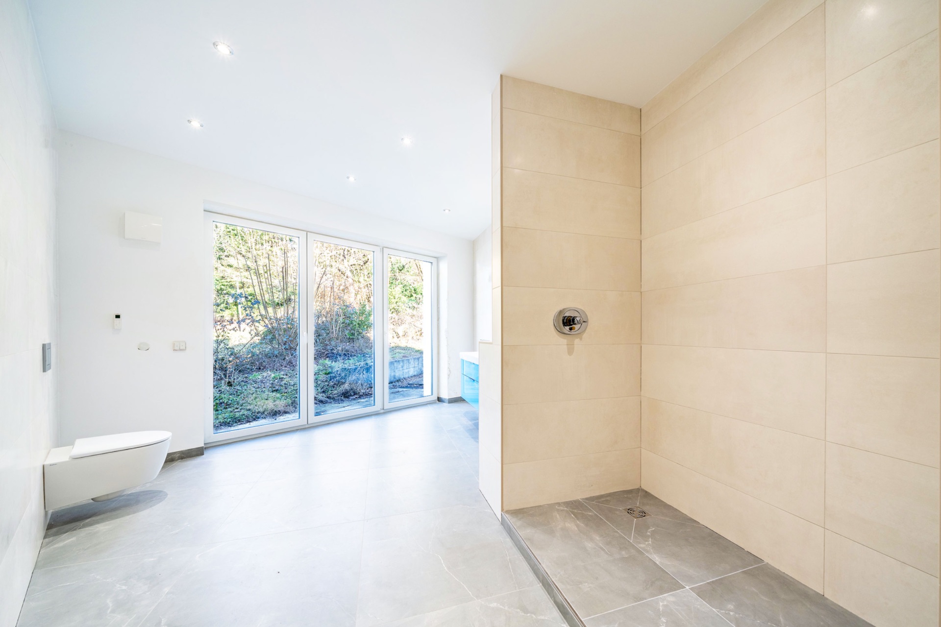 Modernes Badezimmer mit bodengleicher Dusche, WC und großen Fenstern mit Blick ins Grüne.