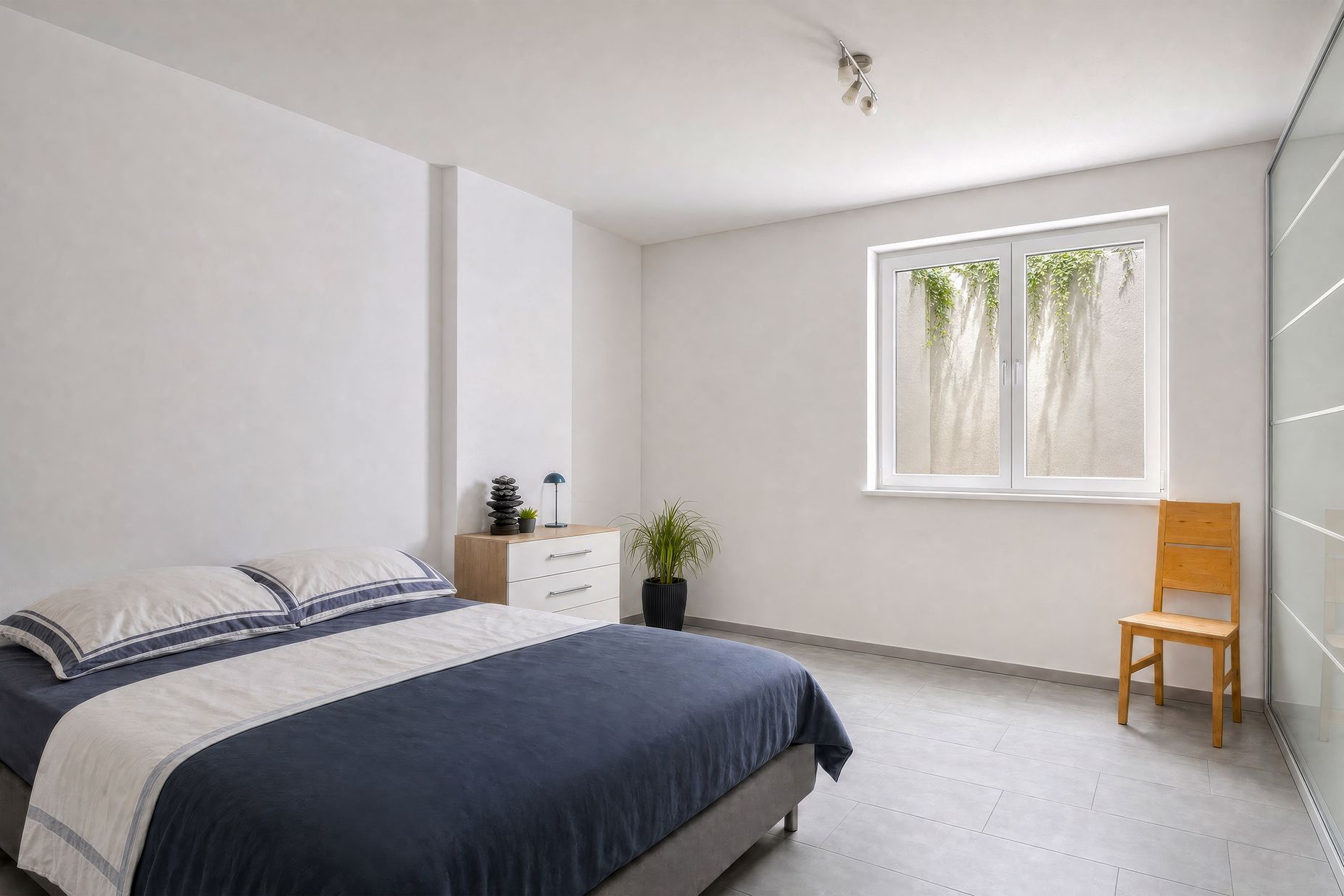 Helles Schlafzimmer mit Doppelbett, Kommode, Stuhl und großem Fenster.