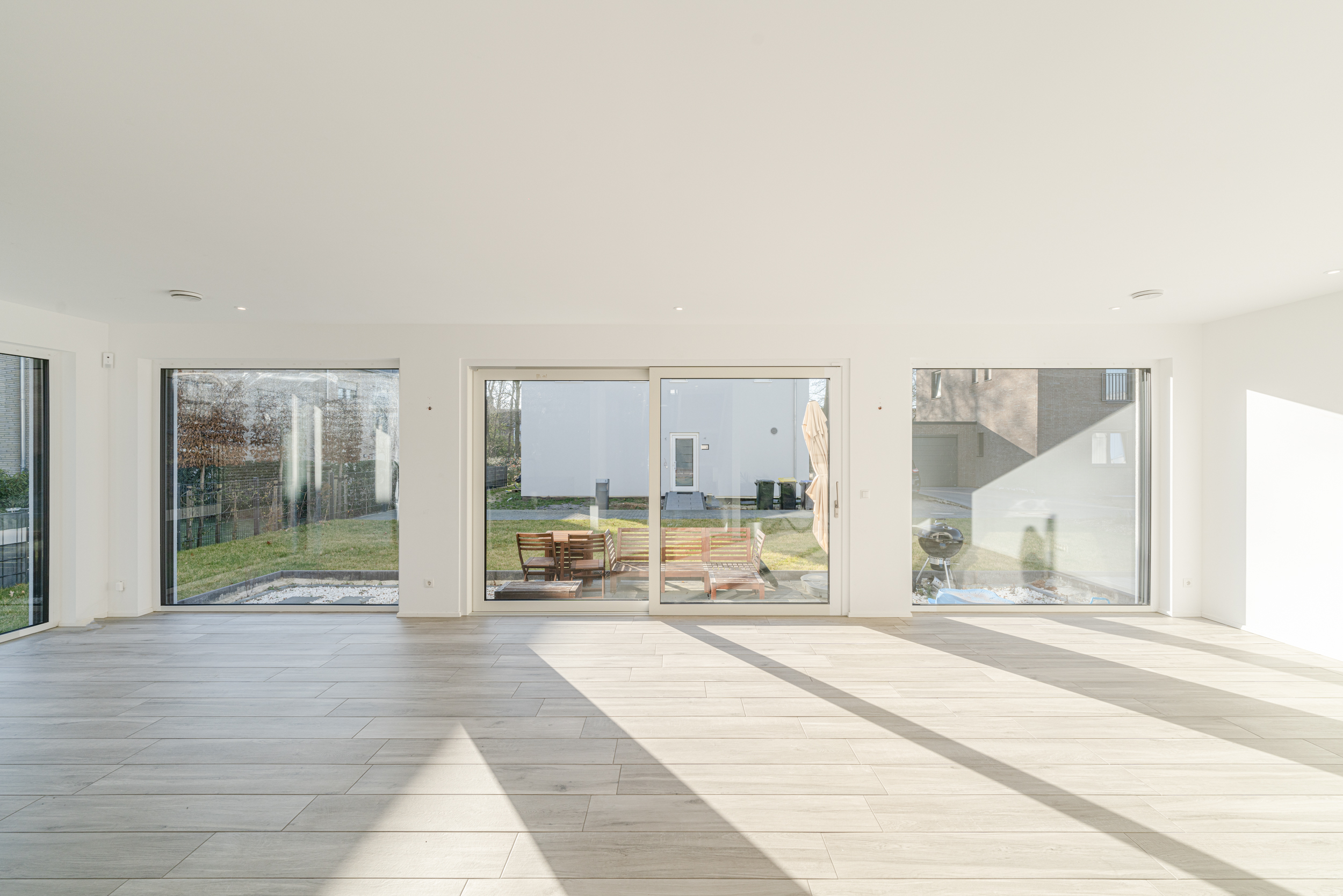 Heller, leerer Raum mit großen Fenstern und Blick auf einen Garten mit Sitzgruppe.
