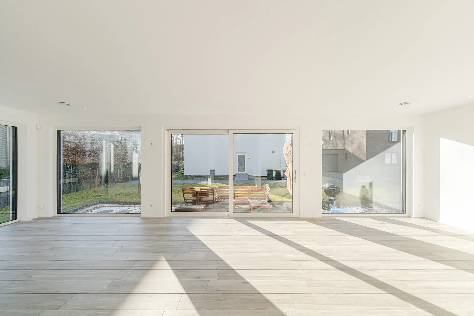 Heller, leerer Raum mit großen Fenstern und Blick auf einen Garten mit Sitzgruppe.