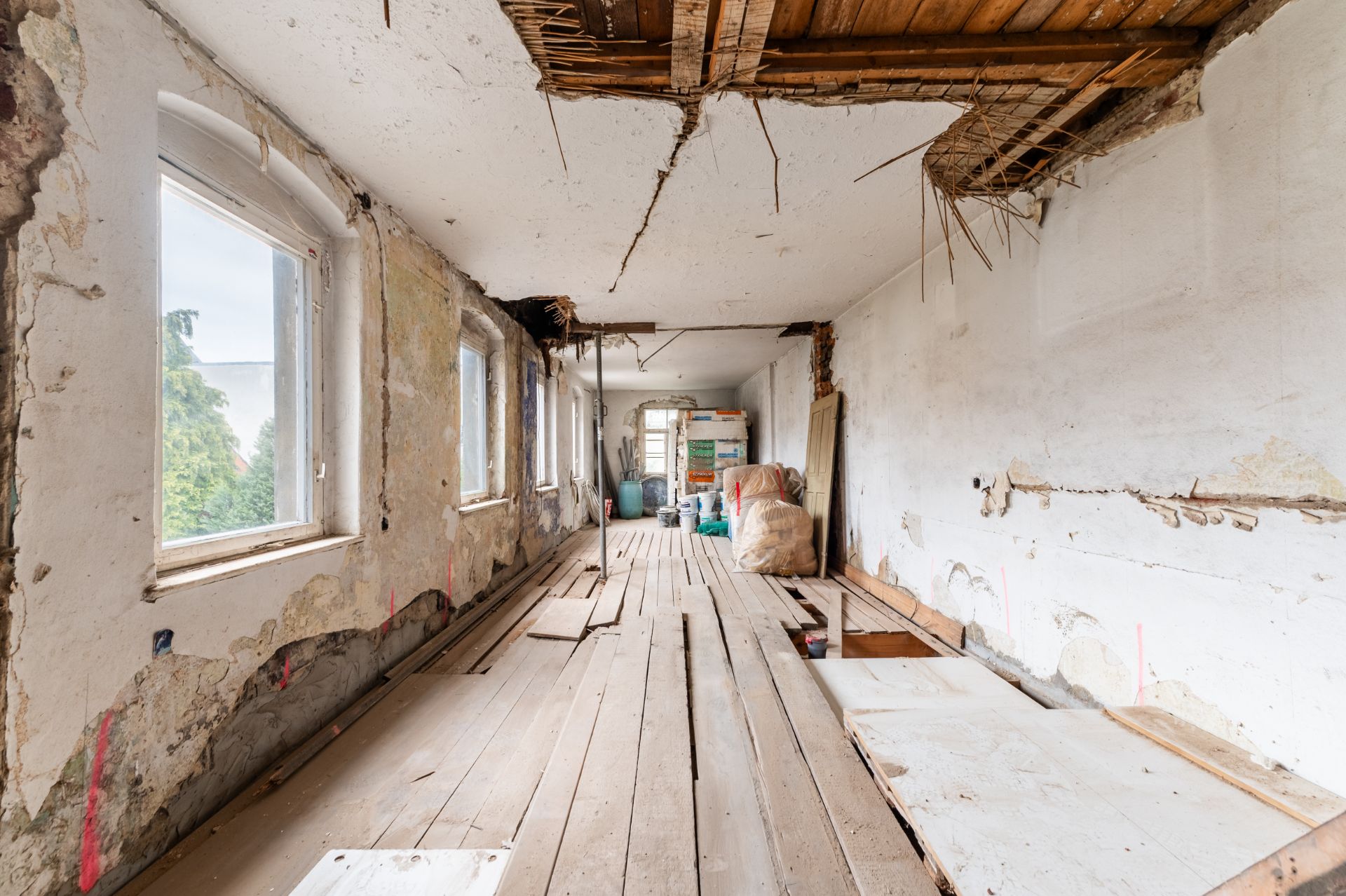Langer Raum mit beschädigten Wänden, offenem Boden und Decke, Renovierungsbedarf erkennbar.