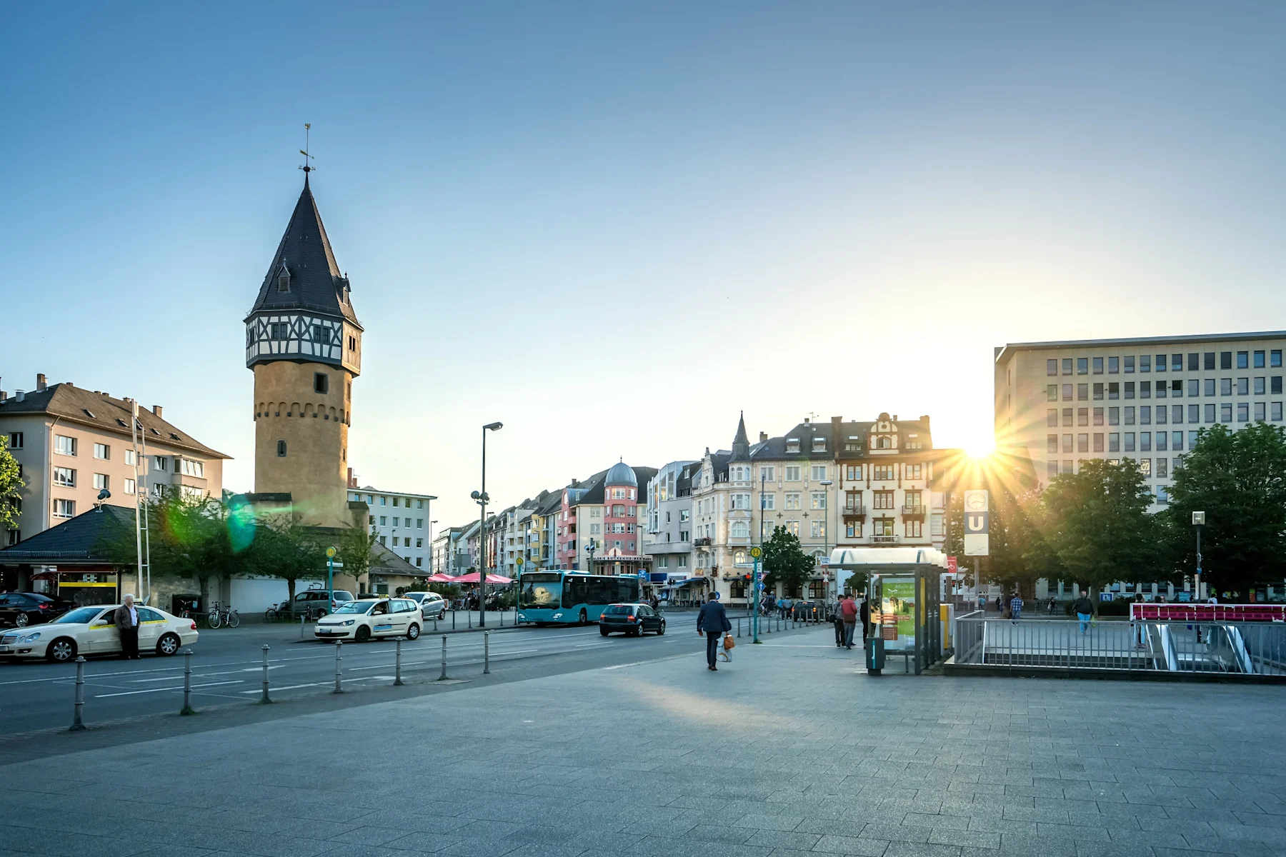 Stadtszene mit historischem Turm, bunten Wohnhäusern, Gehweg und Autos bei Sonnenuntergang.