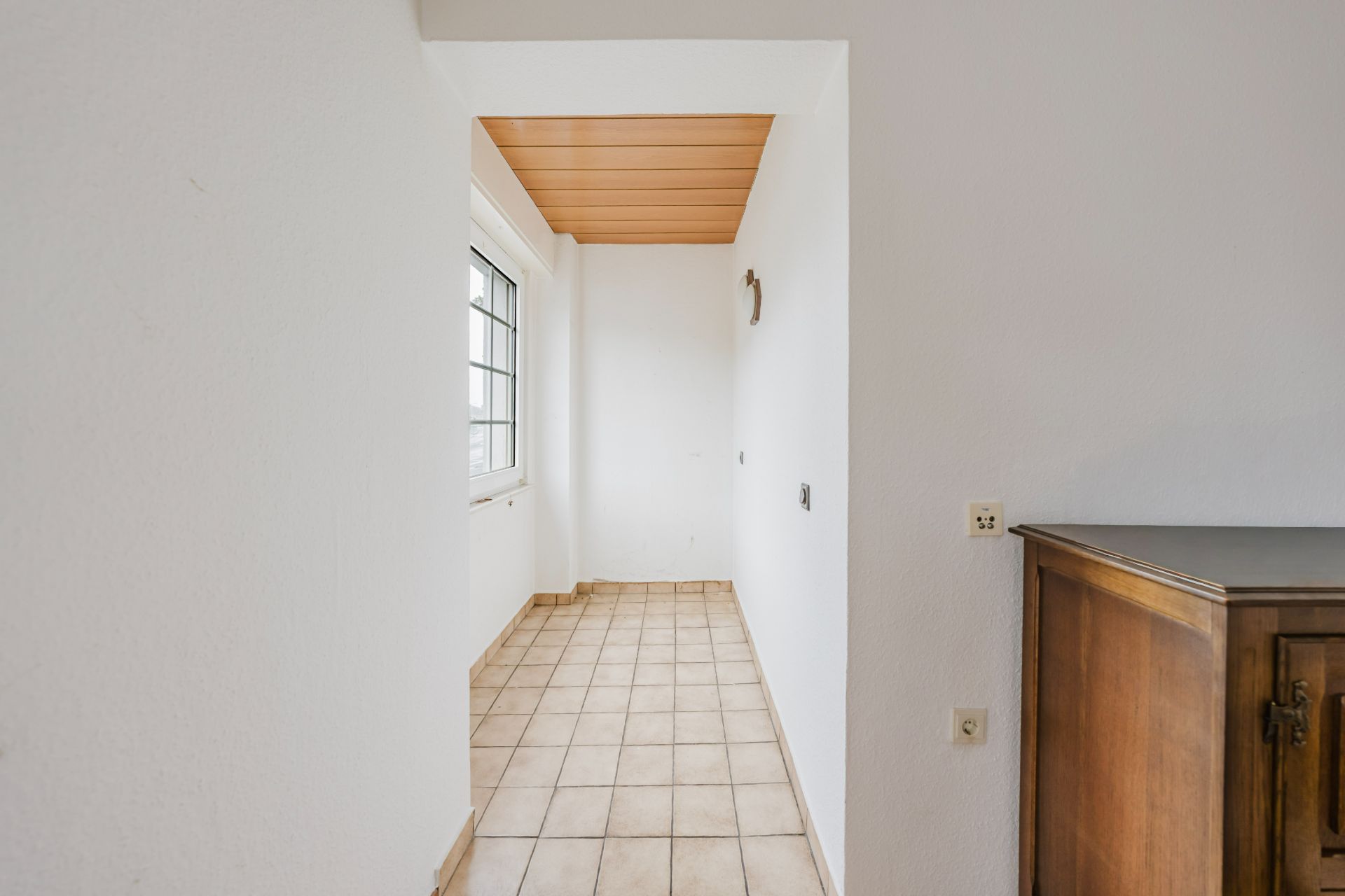 Schmaler Flur mit Fliesenboden, weißer Wand, Fenster und Holzdecke, rechts ein Holzschrank.