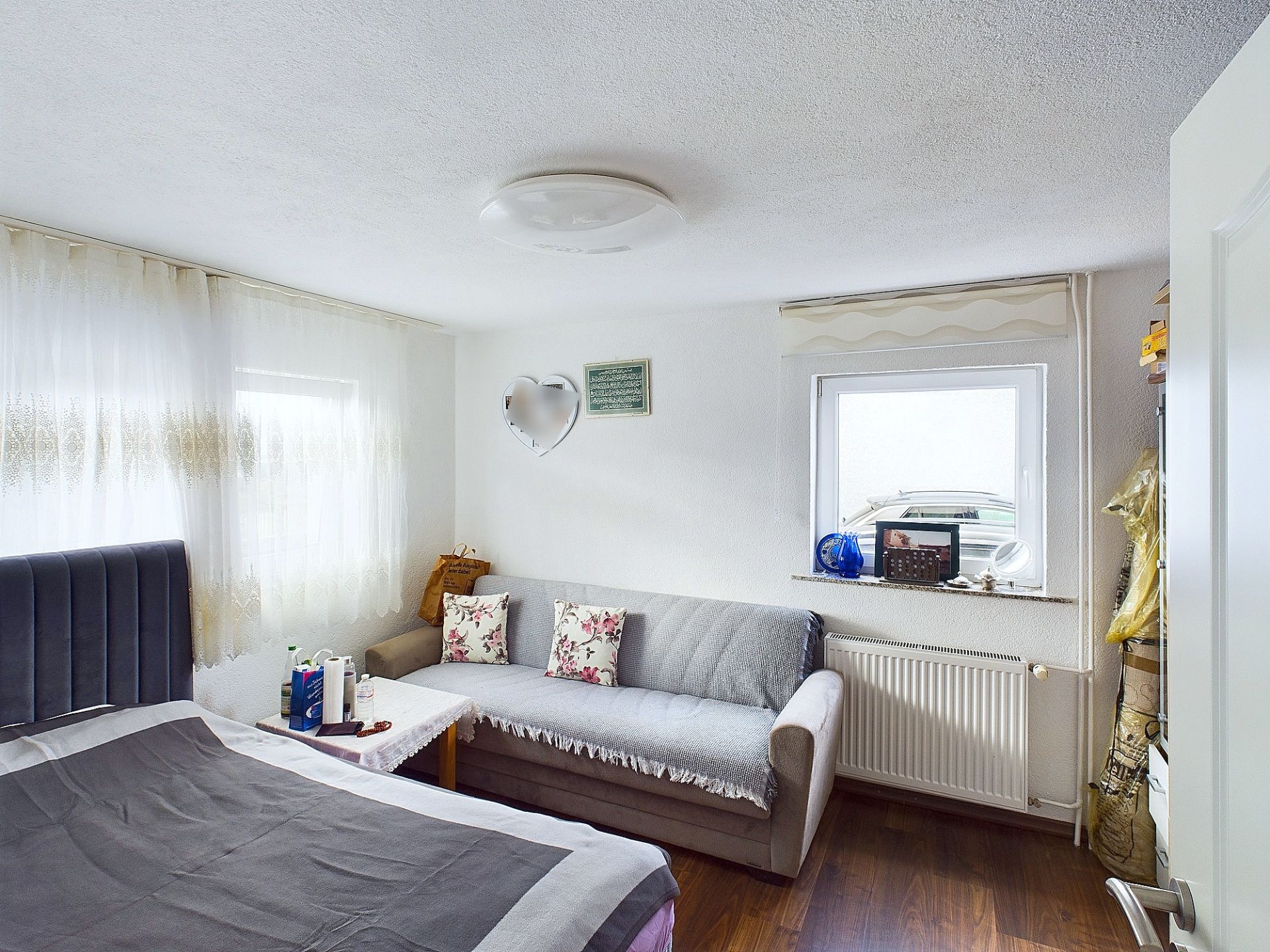 Helles Schlafzimmer mit Bett, Sofa, Tisch und Fenster, dekoriert mit Kissen und Vorhängen.
