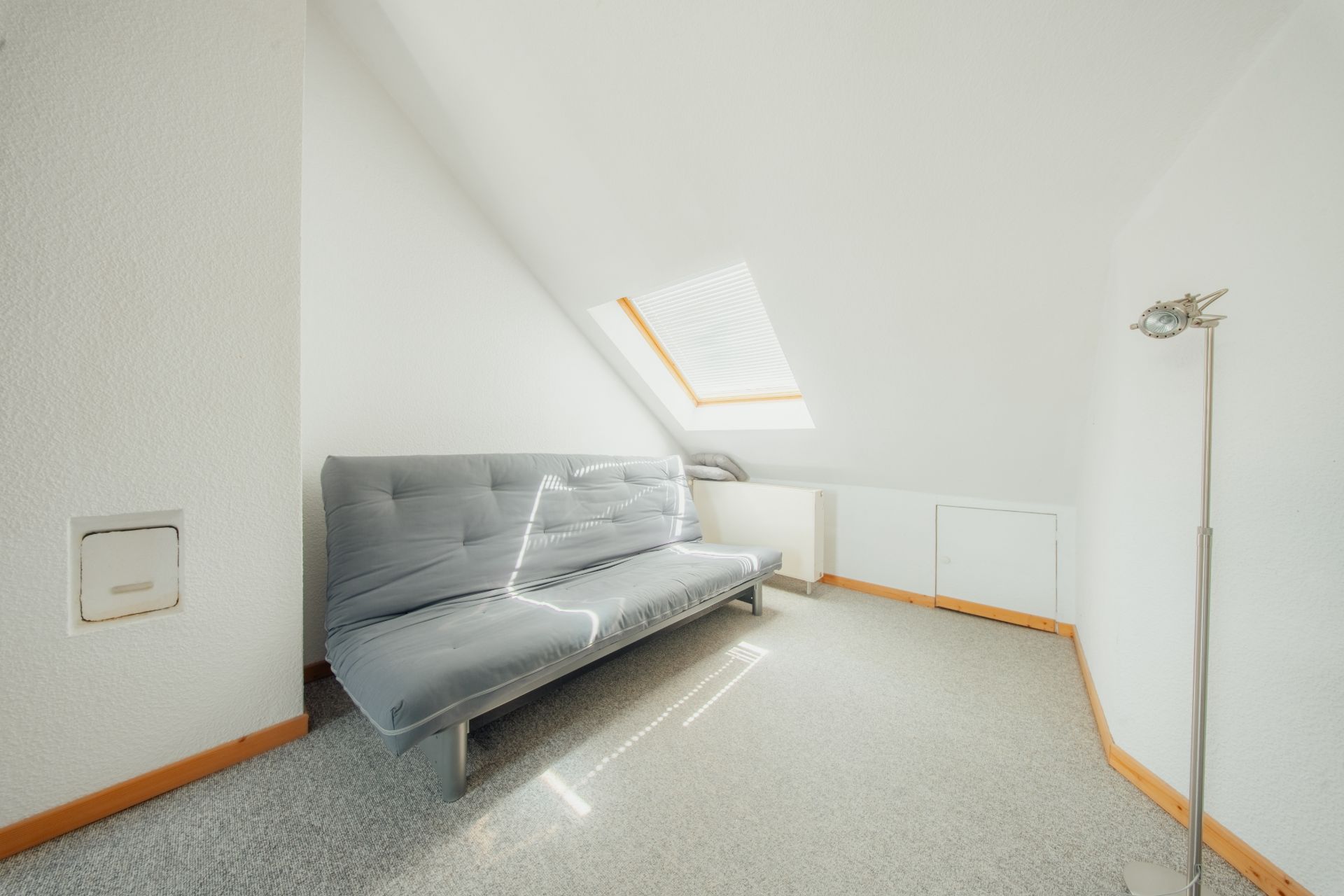 Helles Zimmer mit grauem Sofa, Dachschrägen und Dachfenster, Stehlampe an der Wand.