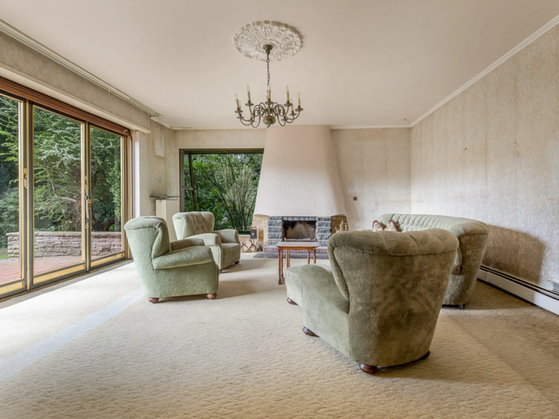 Wohnzimmer mit grünem Sofa, Sesseln, Kamin, Kronleuchter und großen Fenstern zum Garten.