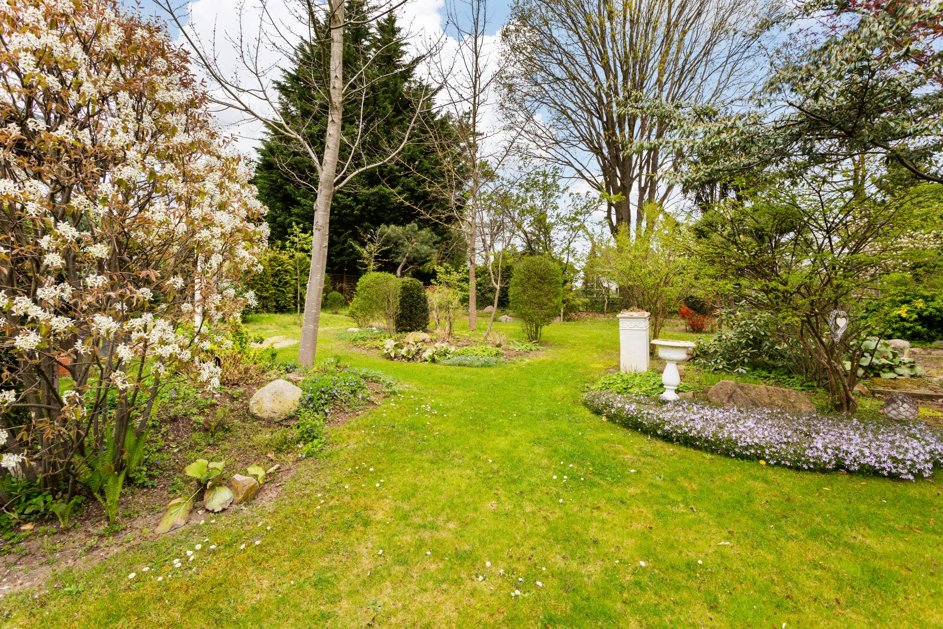 Grüner Garten mit blühenden Büschen, Rasen, Bäumen und einem weißen Vogelbad.