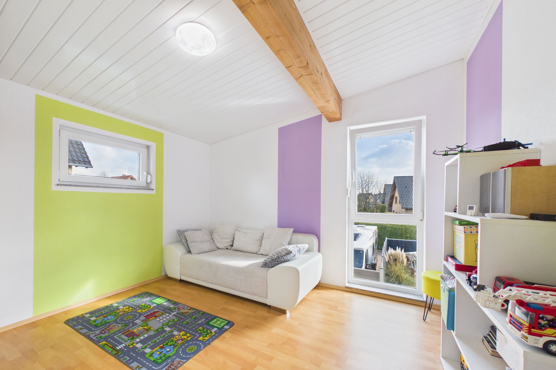 Helles Kinderzimmer mit Sofa, Spielteppich, Regal und großen Fenstern.