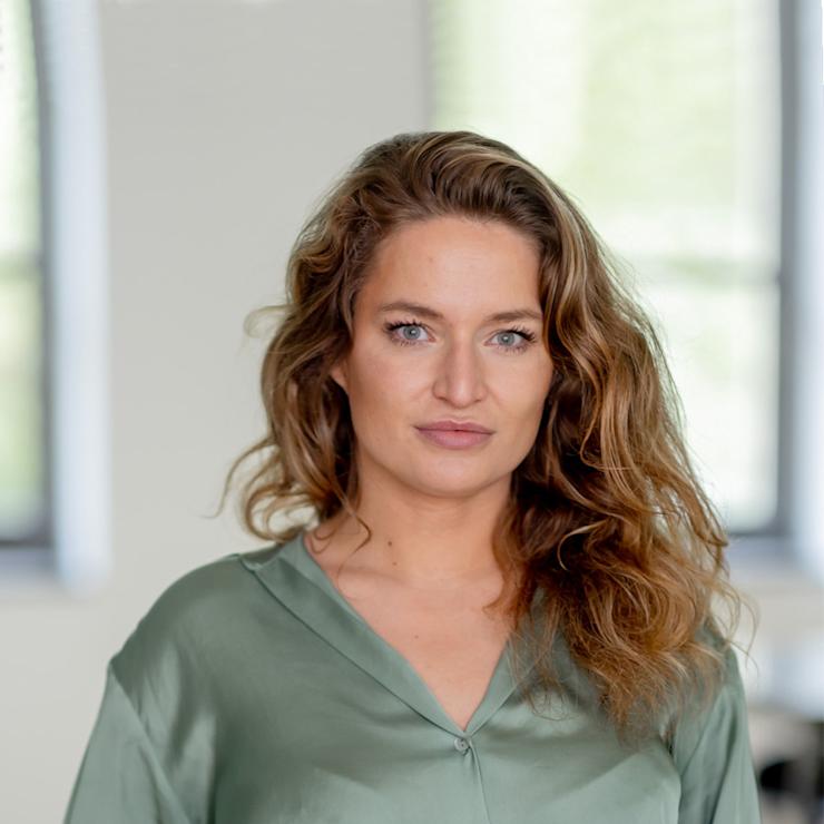 Frau mit langen, lockigen Haaren in grünem Hemd steht in einem hellen Raum.