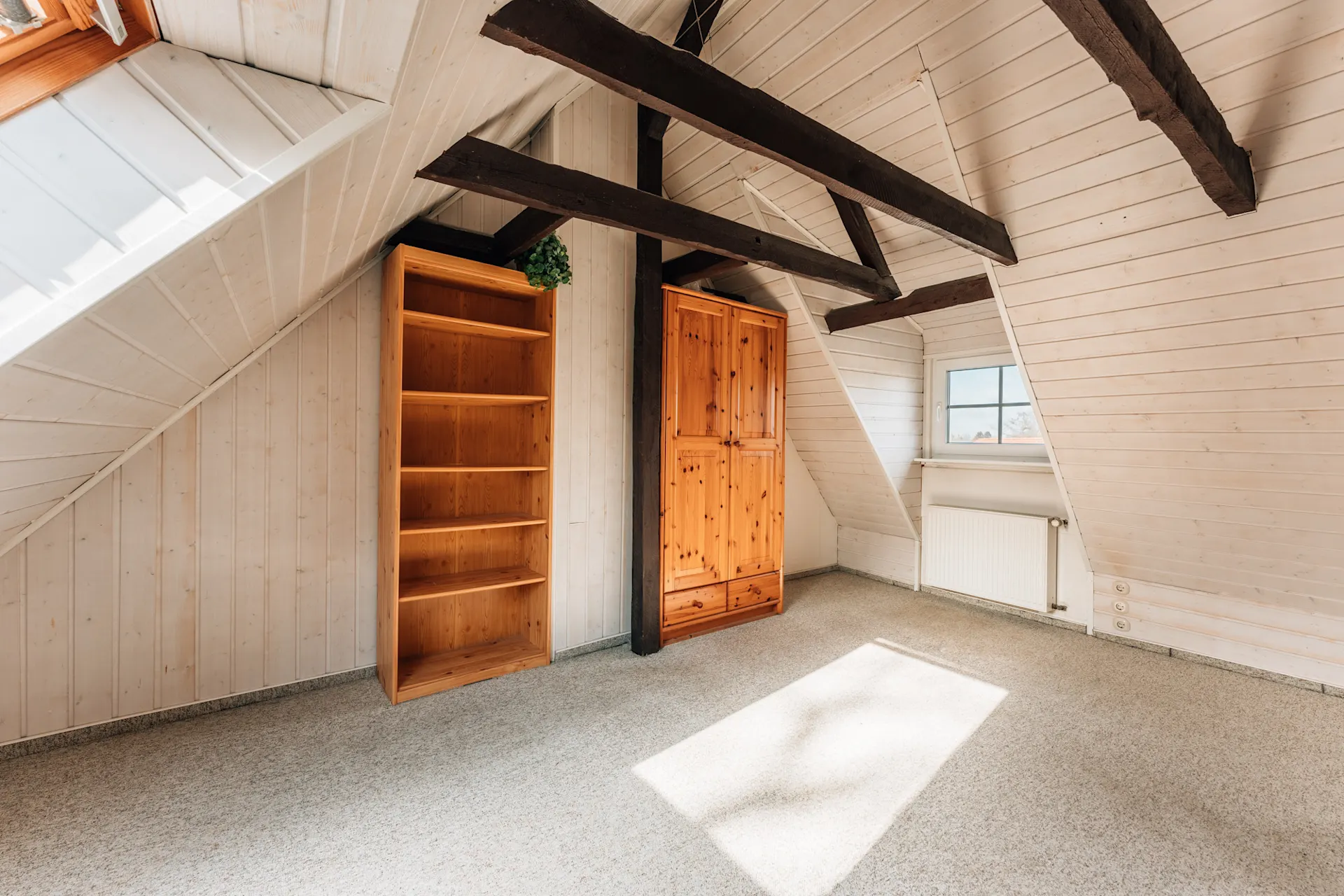 Helles Dachzimmer mit Holzregal, Schrank, Fenster und sichtbaren Holzbalken.