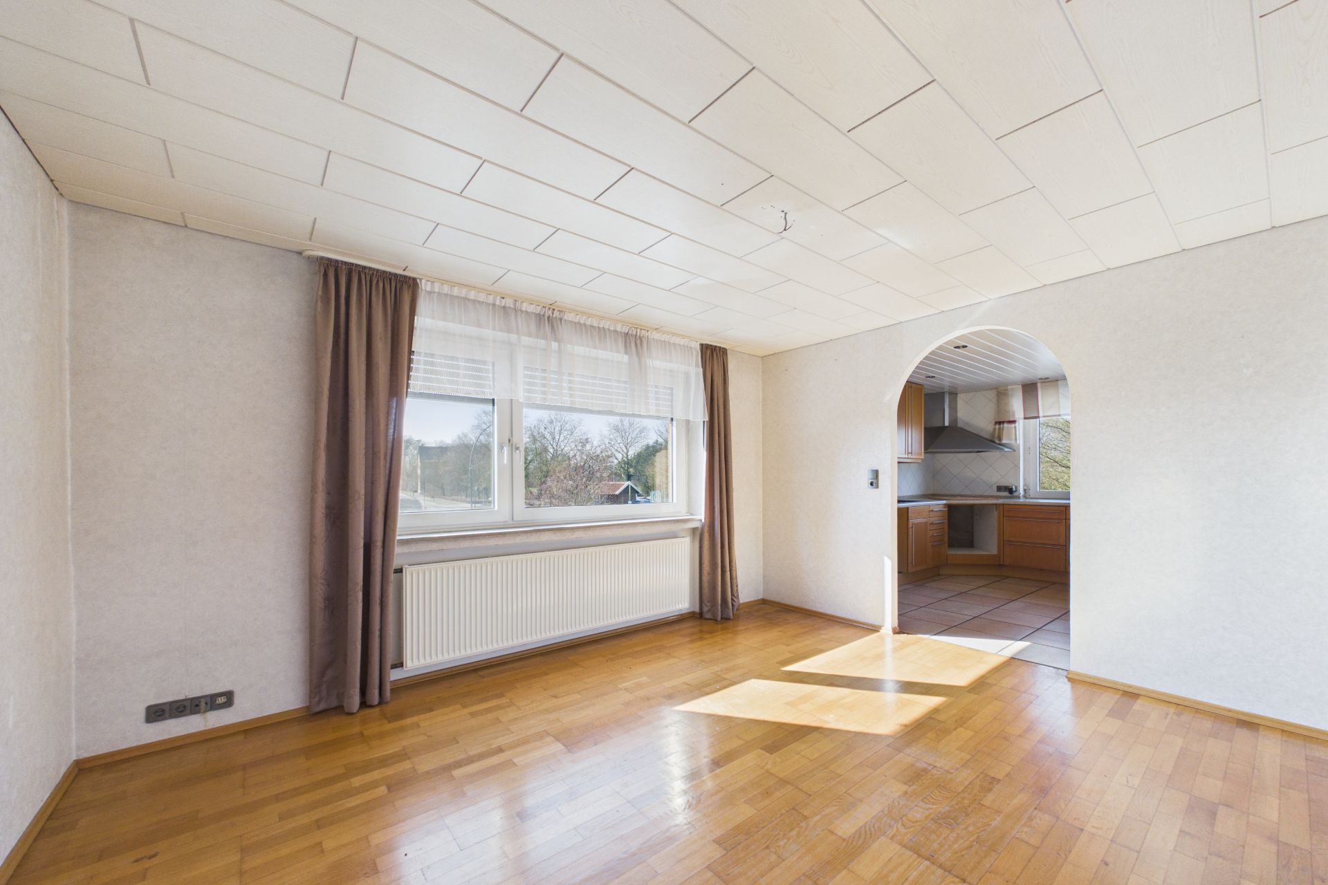 Helles Wohnzimmer mit Parkettboden, Fenster mit Vorhängen und Durchgang zur Küche mit Fliesenboden.