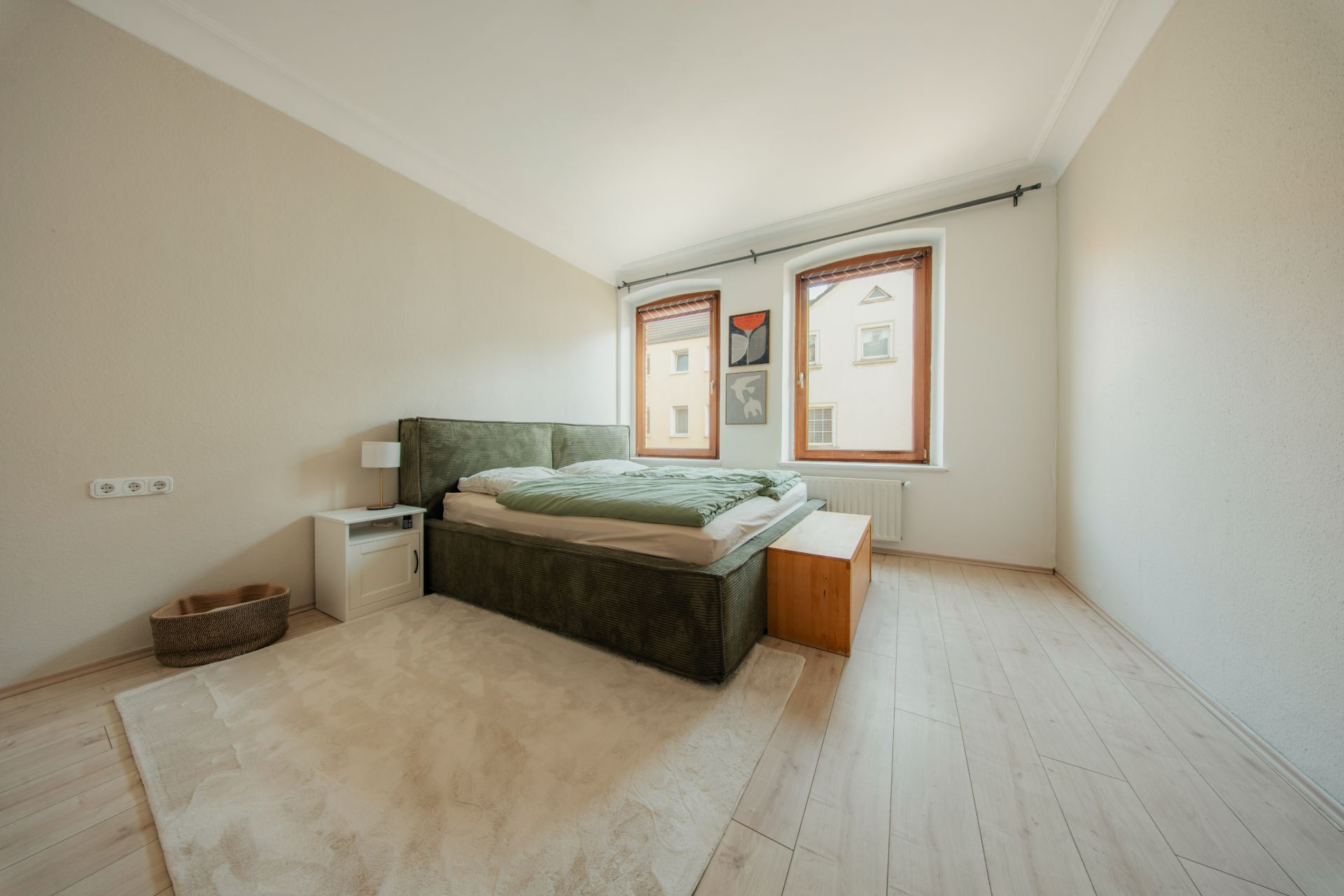 Helles Schlafzimmer mit Doppelbett, Nachttisch, Teppich und zwei großen Fenstern.