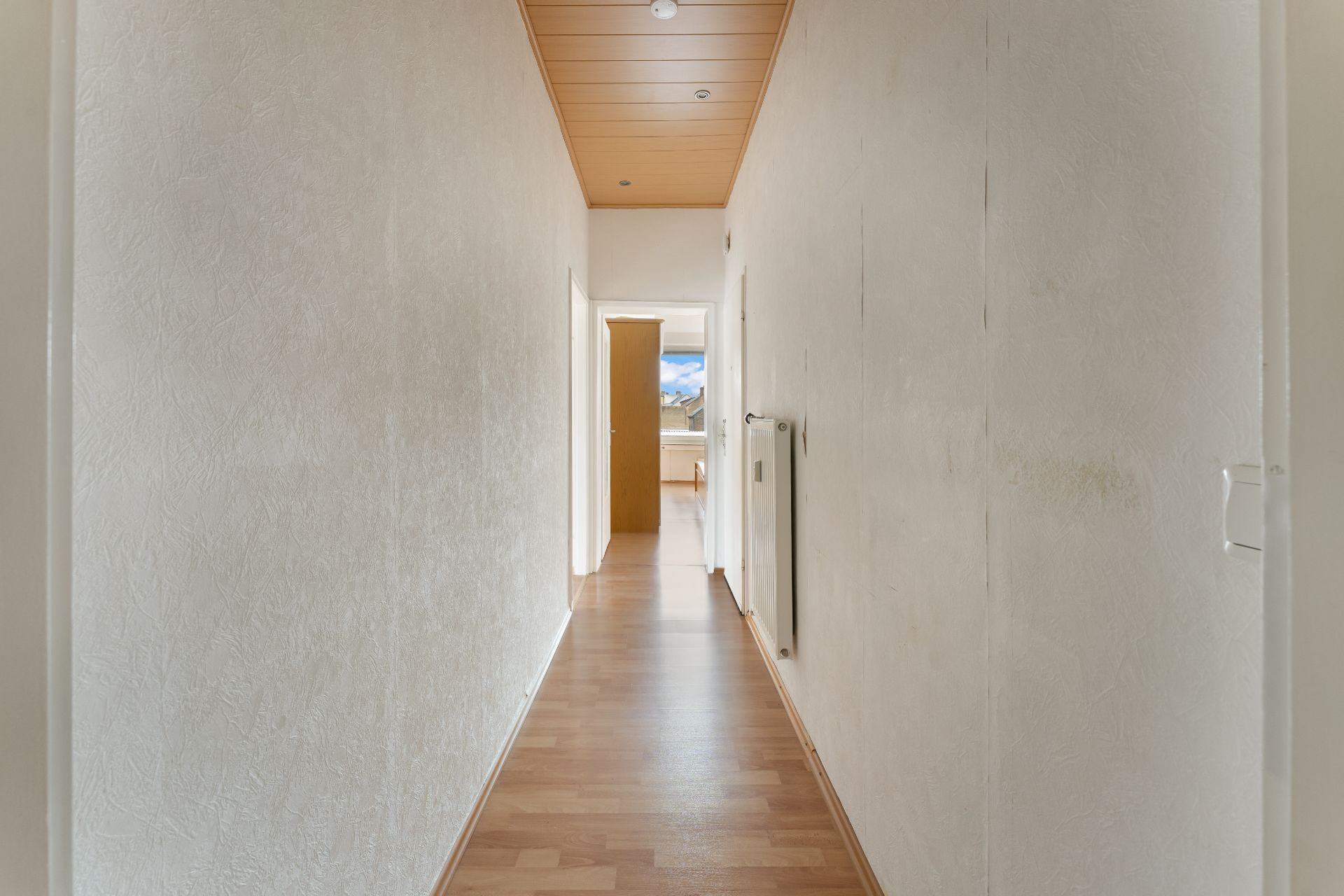 Langer Flur mit Holzfußboden, weiß gestrichenen Wänden und einer Heizung an der rechten Wand.