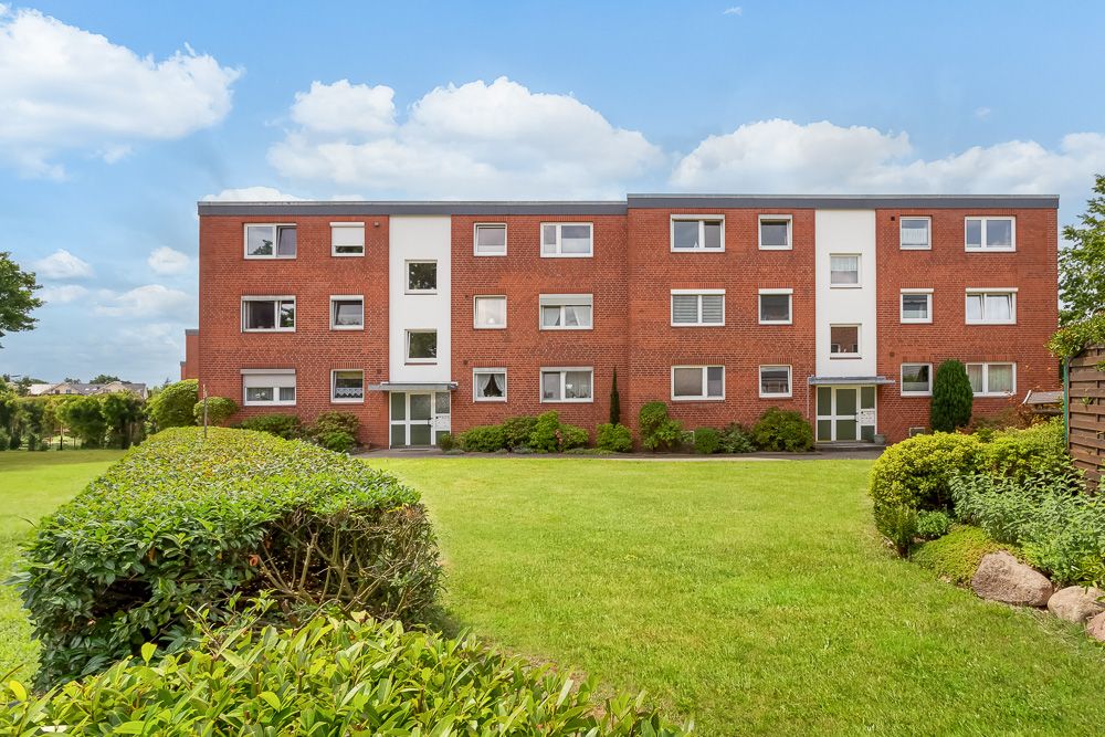 Rotes Mehrfamilienhaus mit gepflegtem Vorgarten und grüner Hecke, drei Stockwerke, viele Fenster.