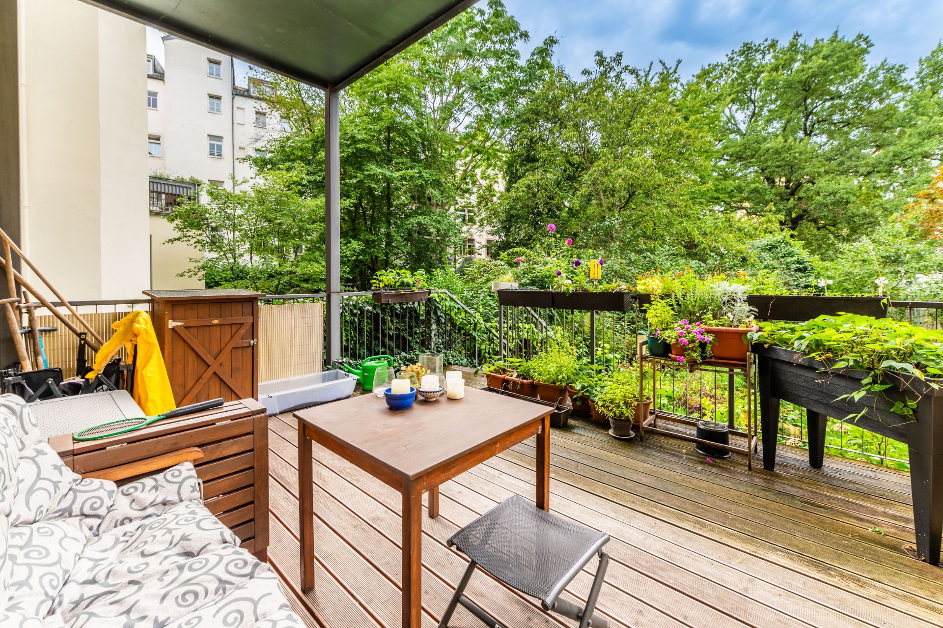 Überdachte Terrasse mit Holzboden, Holztisch, Stuhl, Sofa und vielen Pflanzen am Geländer.
