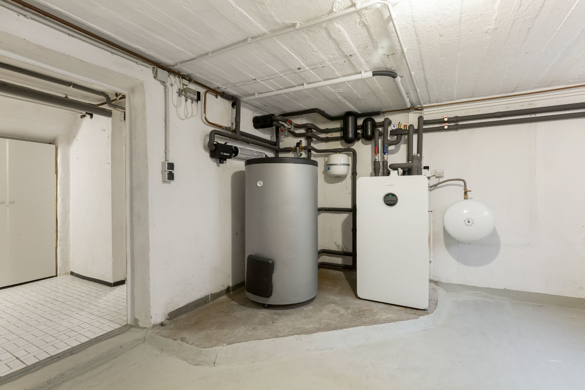 Heizungsanlage mit Rohren und Tanks in einem Kellerraum, weiße Wände und grauer Boden.
