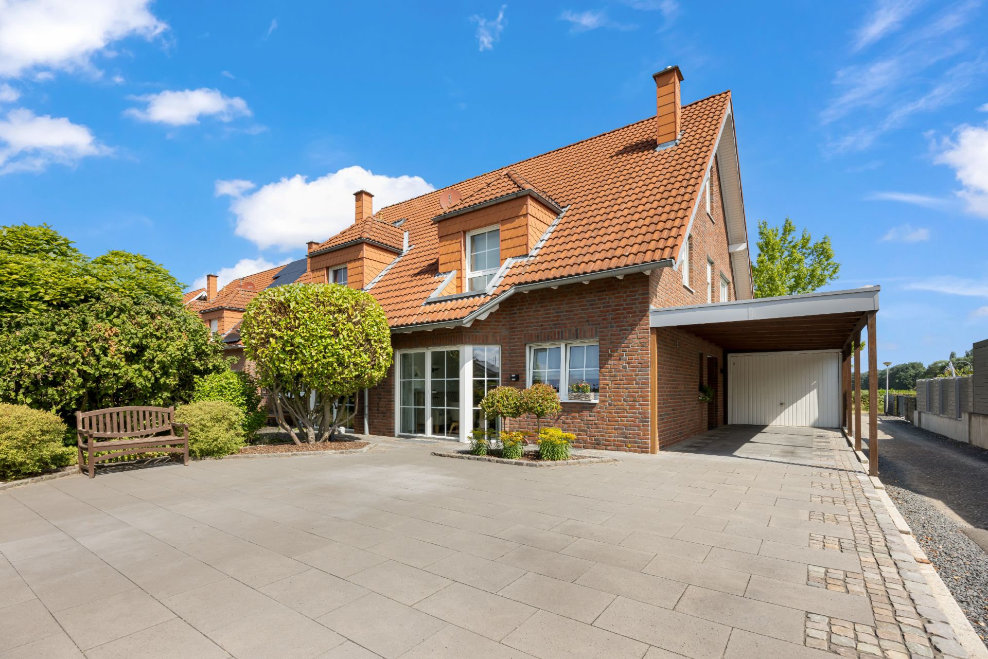 Zweistöckiges Backsteinhaus mit rotem Dach, großer Einfahrt, Carport und Gartenbank.