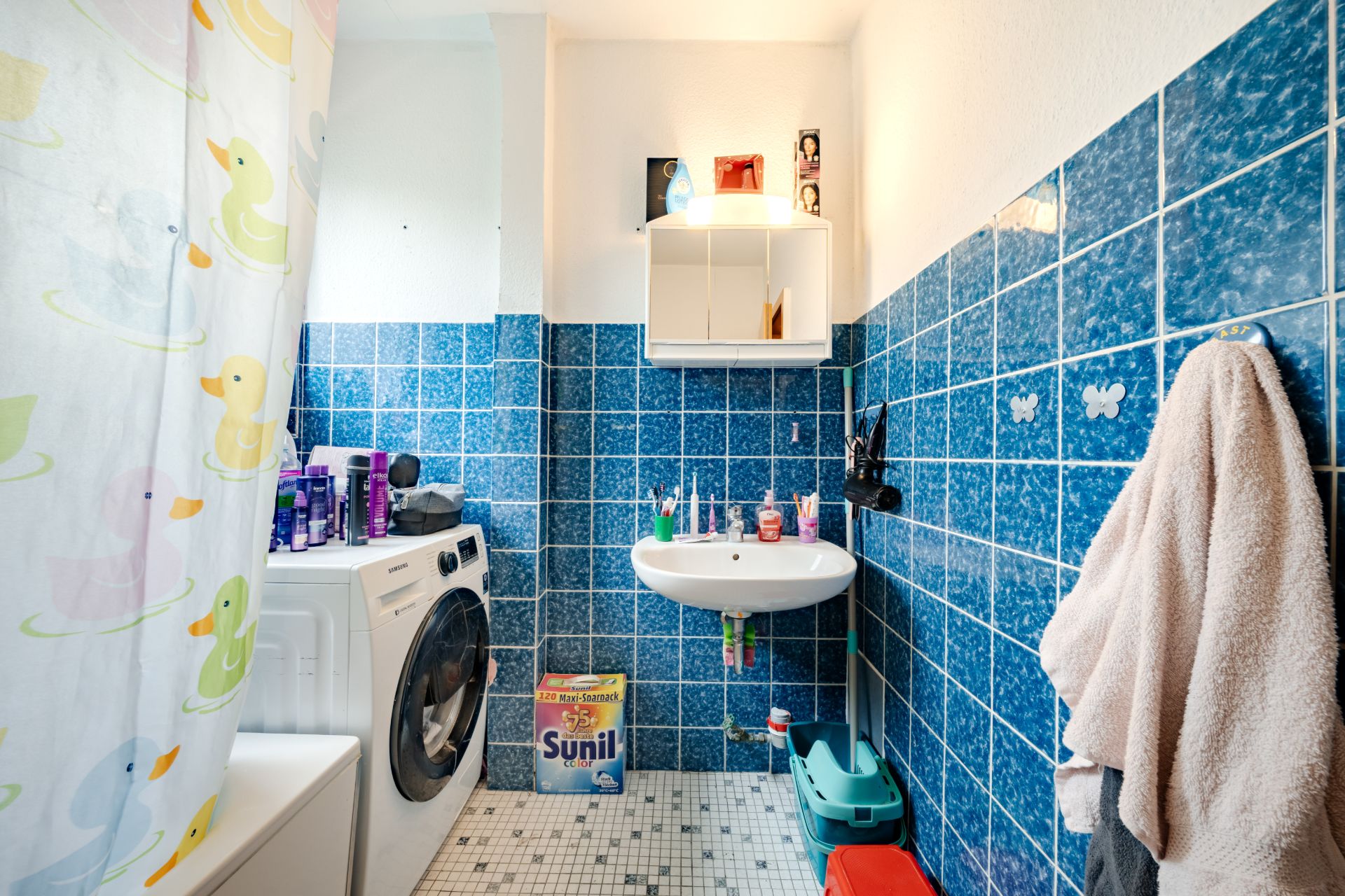 Badezimmer mit blauen Fliesen, Waschmaschine, Waschbecken, Duschvorhang mit Enten, Handtuch und Putzmitteln.