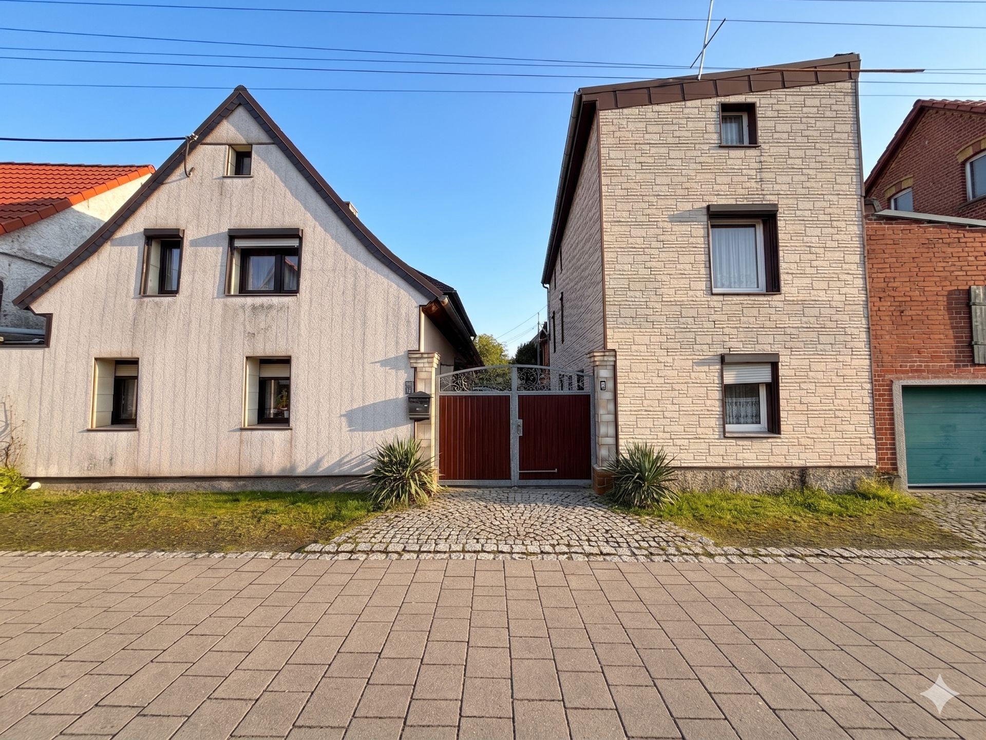 Zwei Wohnhäuser mit Tor und Pflastersteinen vor dem Eingang, einige Pflanzen daneben.