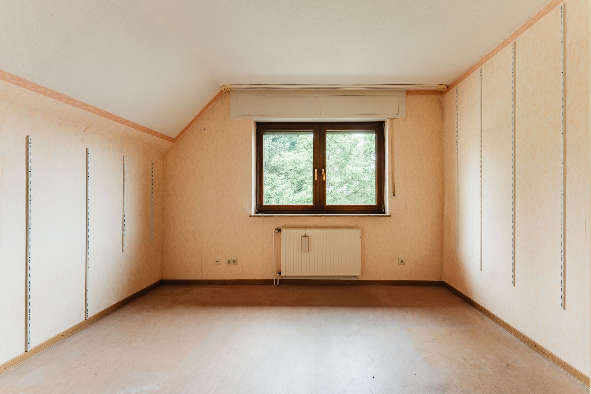 Leeres Zimmer mit Dachschräge, Fenster und Heizkörper, helle Wände mit Schienen an den Seiten.