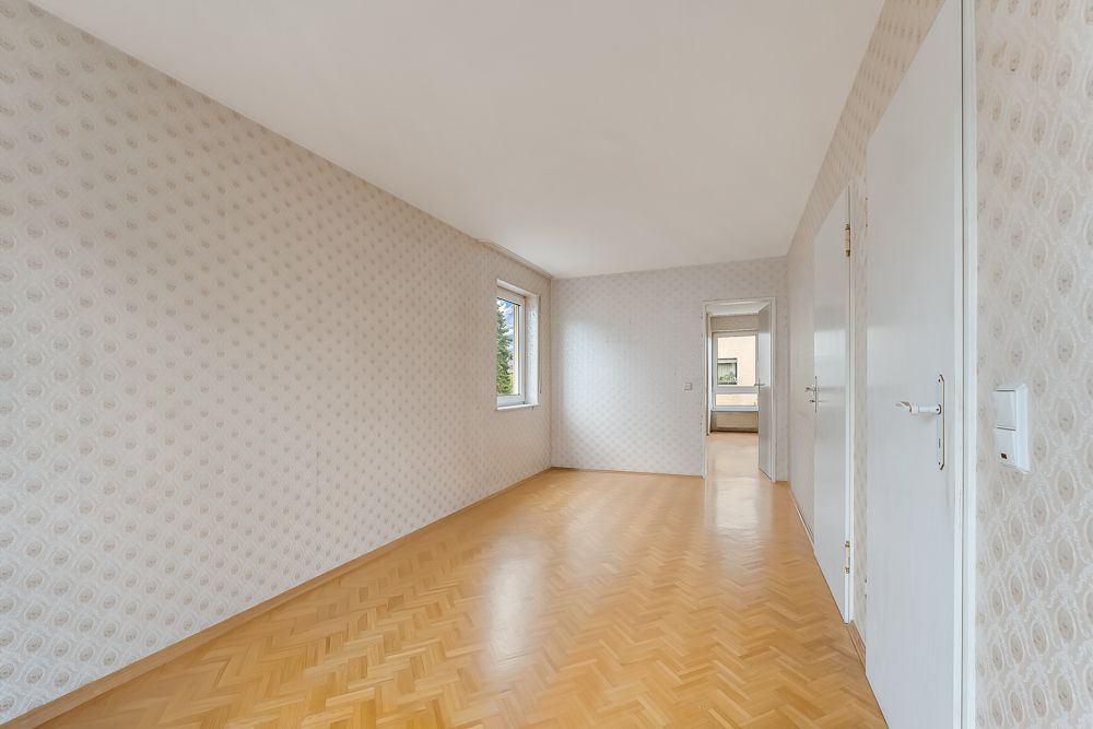 Langer, leerer Raum mit Parkettboden, gemusterten Tapeten und zwei Fenstern.