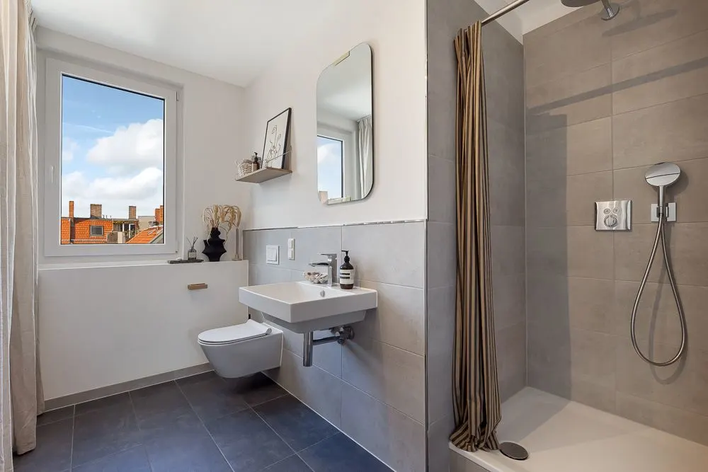 Modernes Badezimmer mit Fenster, Waschbecken, Spiegel, WC und offener Dusche mit grauen Fliesen.