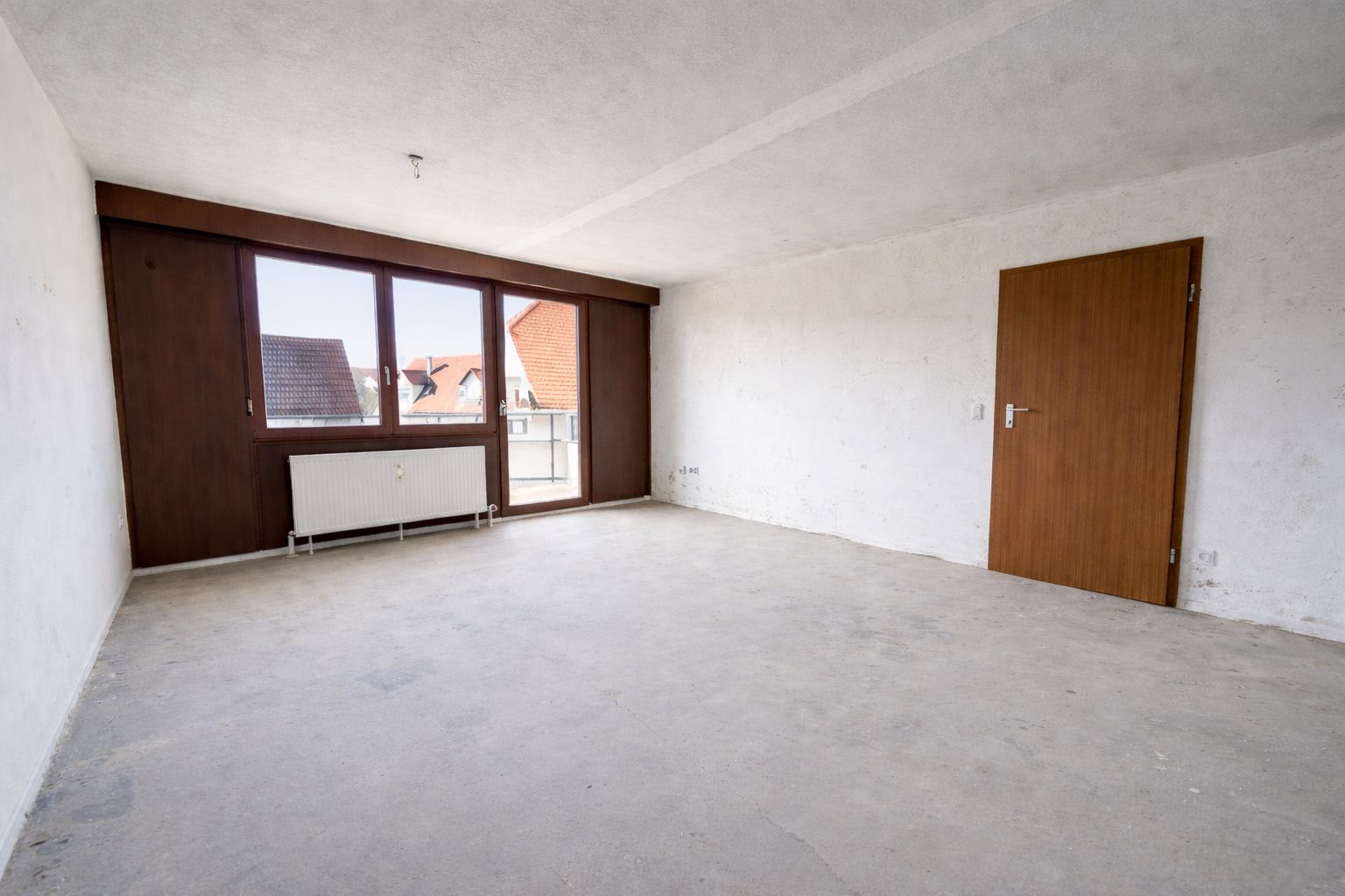 Leerer Raum mit großen Fenstern, weißem Boden, Holztür und Heizkörper unter dem Fenster.