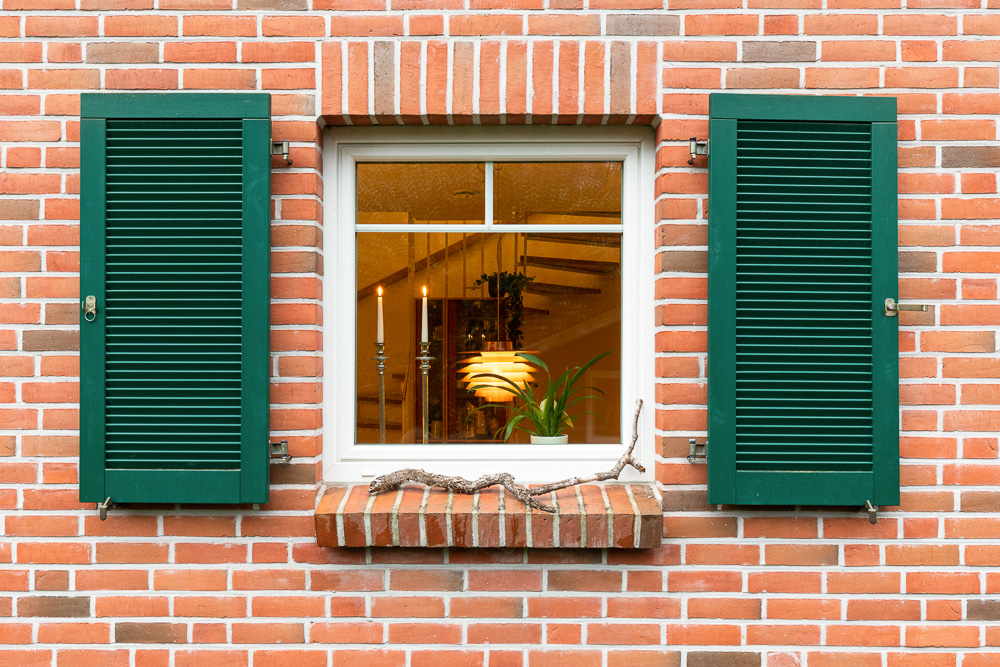Backsteinfassade mit Fenster, grünen Fensterläden und Kerzen im Inneren sichtbar.