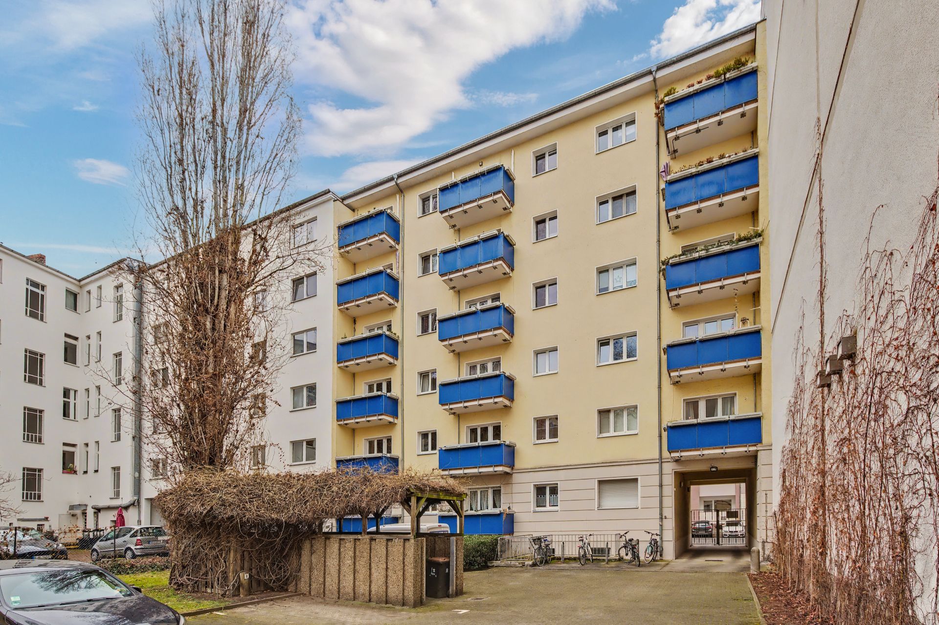 Mehrfamilienhaus mit gelber Fassade und blauen Balkonen, Innenhof mit Mülltonnen und Fahrrädern.
